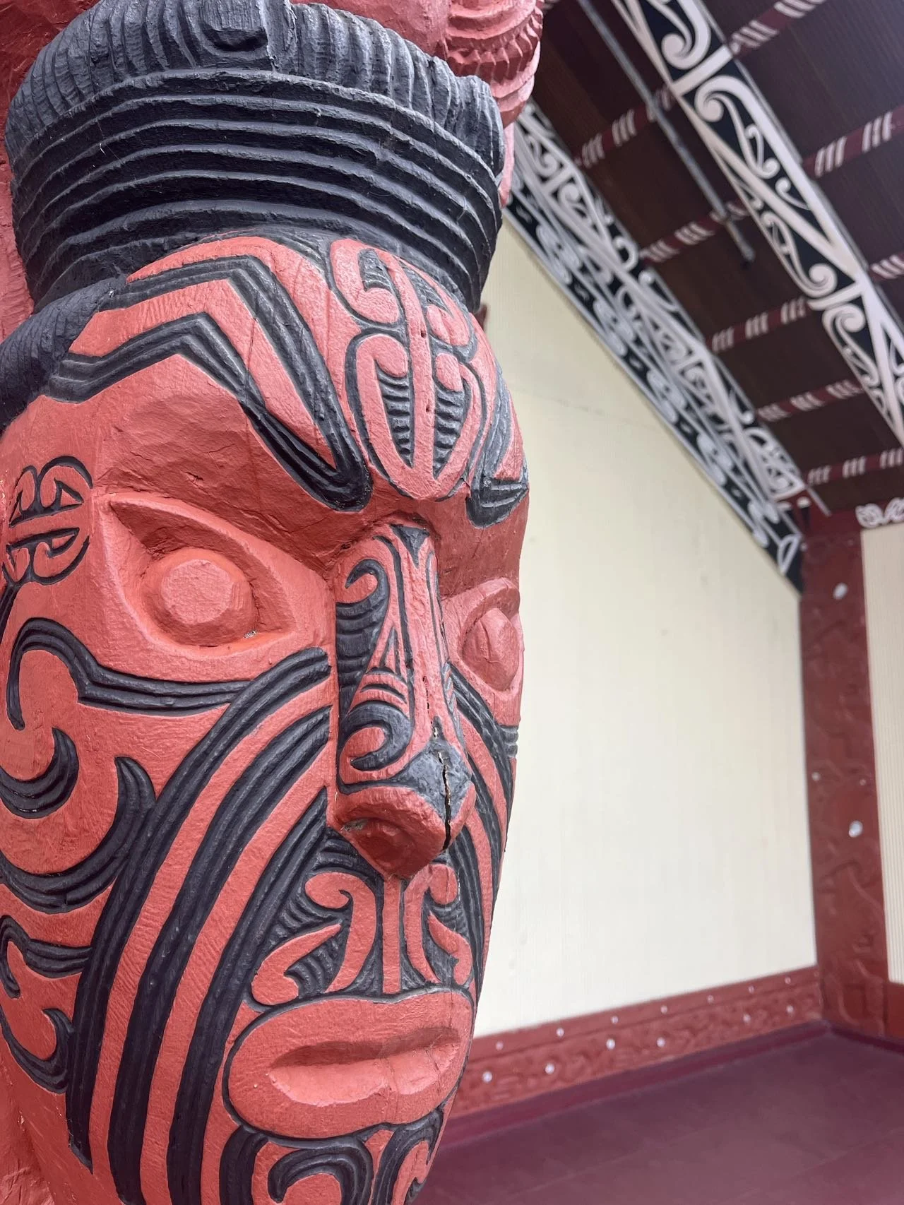 A close-up of a traditional Māori wooden carving featuring intricate black facial moko (tattoo) patterns on a red-painted face, set against the backdrop of a meeting house with painted kōwhaiwhai ceiling rafters.