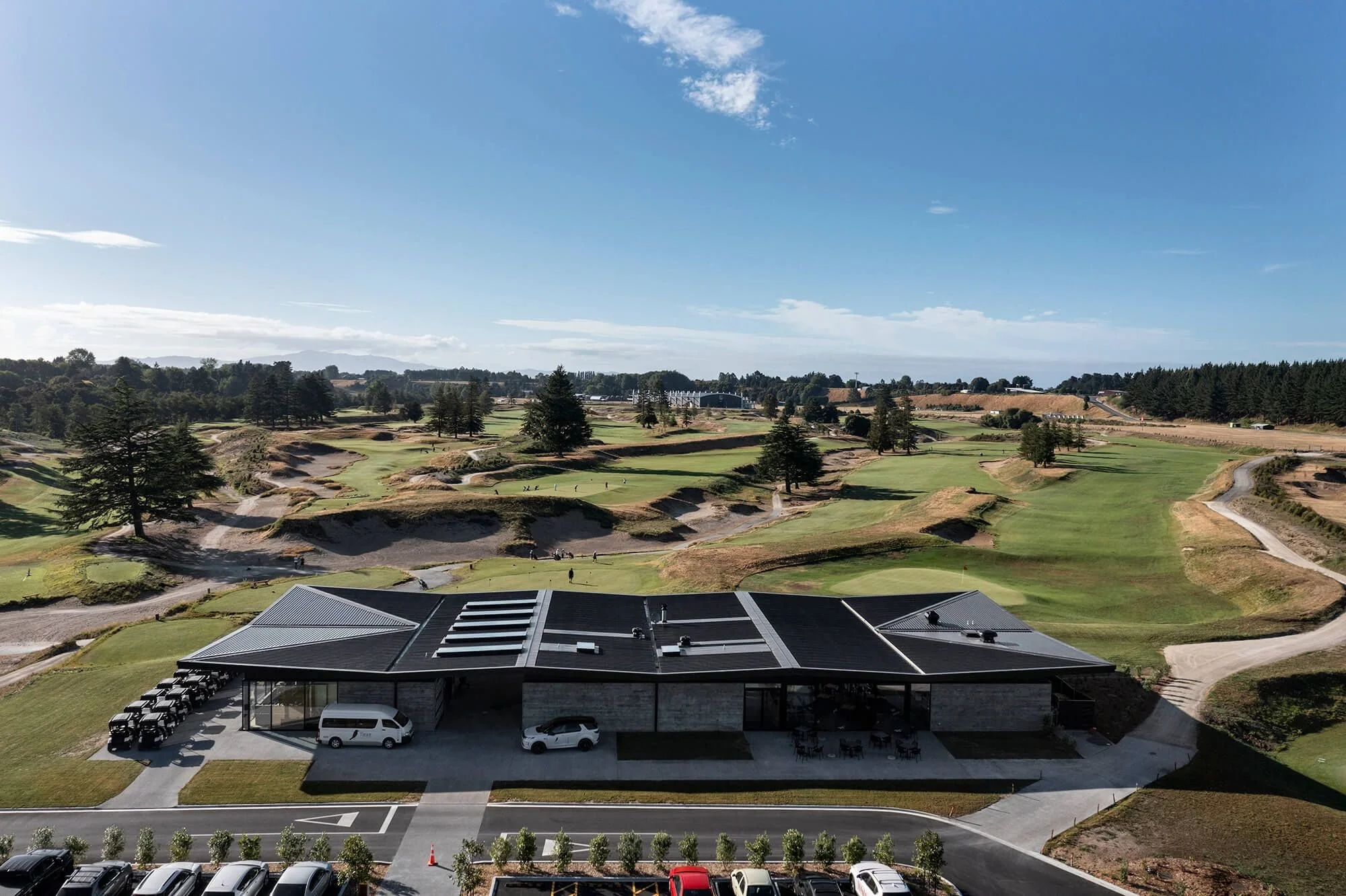 Aerial view of a golf course with a clubhouse in the foreground, surrounded by landscaped greens, trees, and sand bunkers under a clear blue sky.