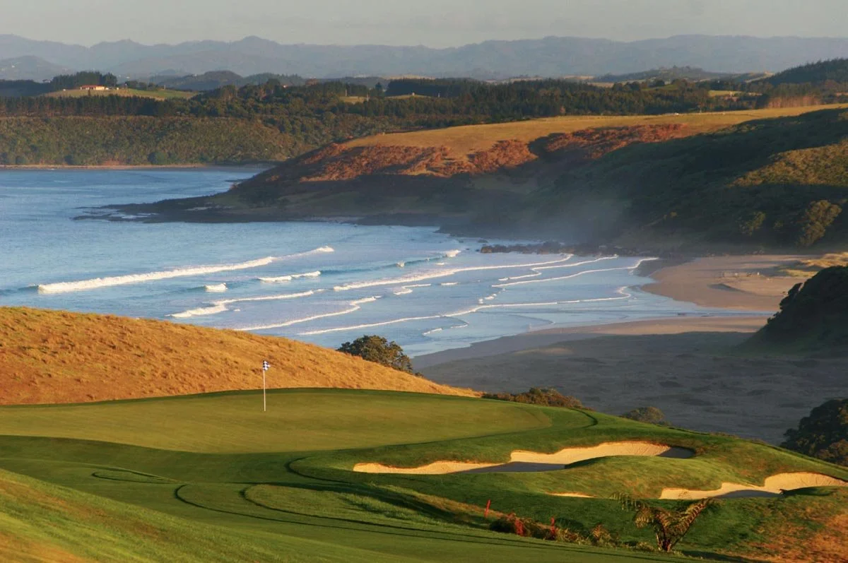 Scenic coastal golf course overlooking a beach, with rolling green hills, sand bunkers, and ocean waves in the background.