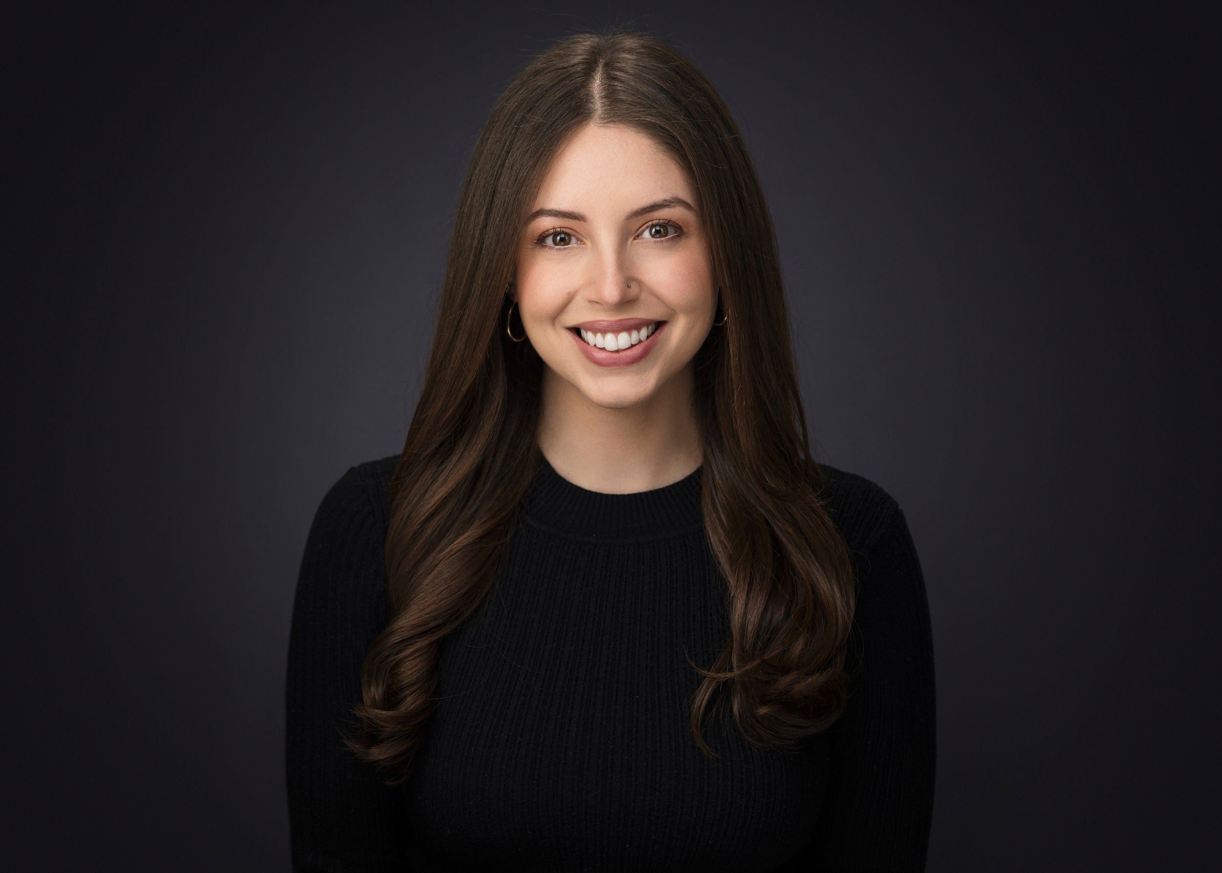Headshot of a young woman with long brown hair, smiling, wearing a black top, against a dark gray background.