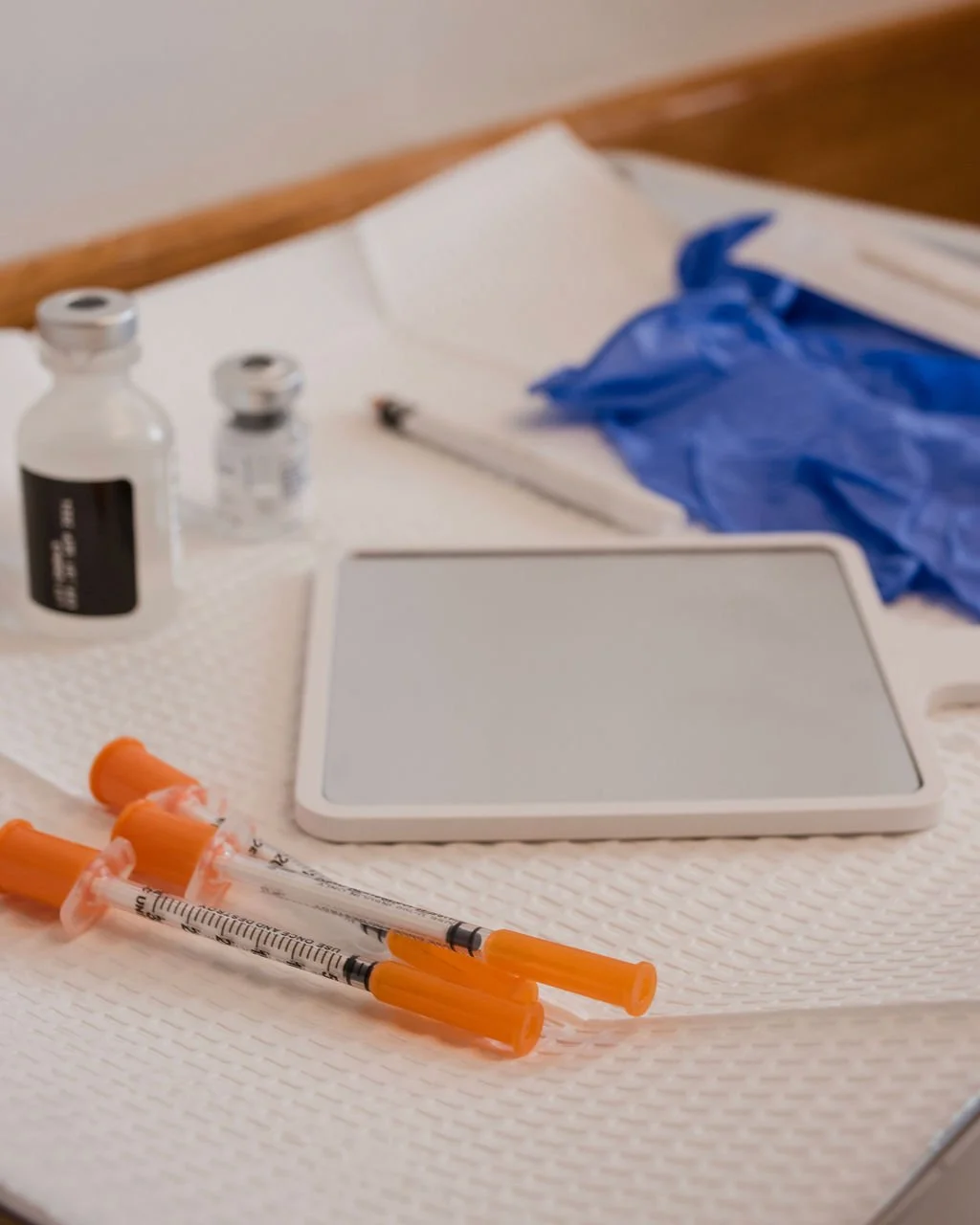 Medical supplies including syringes with orange caps, botox vials, a mirror, a pair of blue gloves, and a small bottle on a white surface.