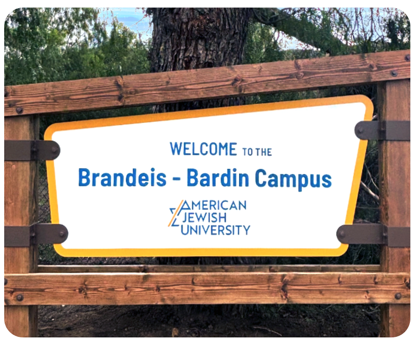 Welcome sign for Brandeis Bardin Campus of American Jewish University, mounted on a wooden frame outdoors with trees in the background.
