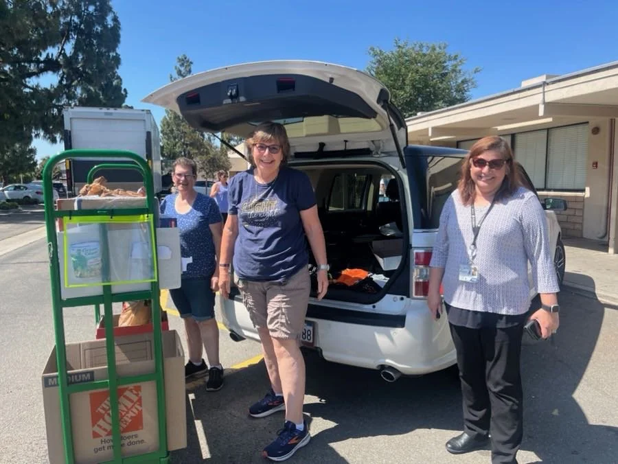 Three women standing outside a white SUV with the back open, in a parking lot on a sunny day. One woman is holding a phone, another is leaning on the car, and the third is standing behind a green cart with food items and boxes.