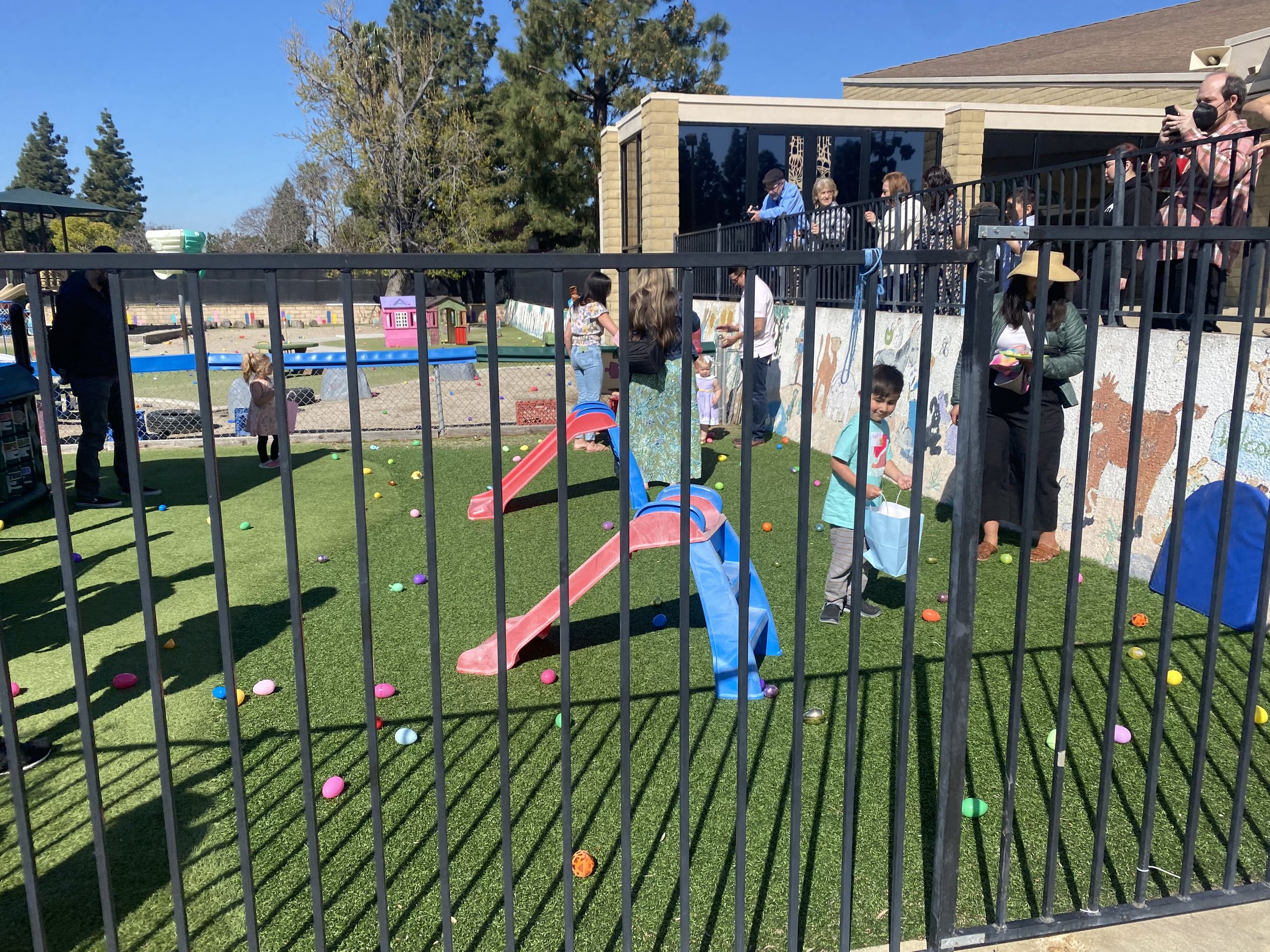 Children and adults gather around a fenced area with a small slide, collecting colorful plastic eggs on artificial grass during an Easter egg hunt. There are onlookers on an upper balcony, and a mural on the wall behind.