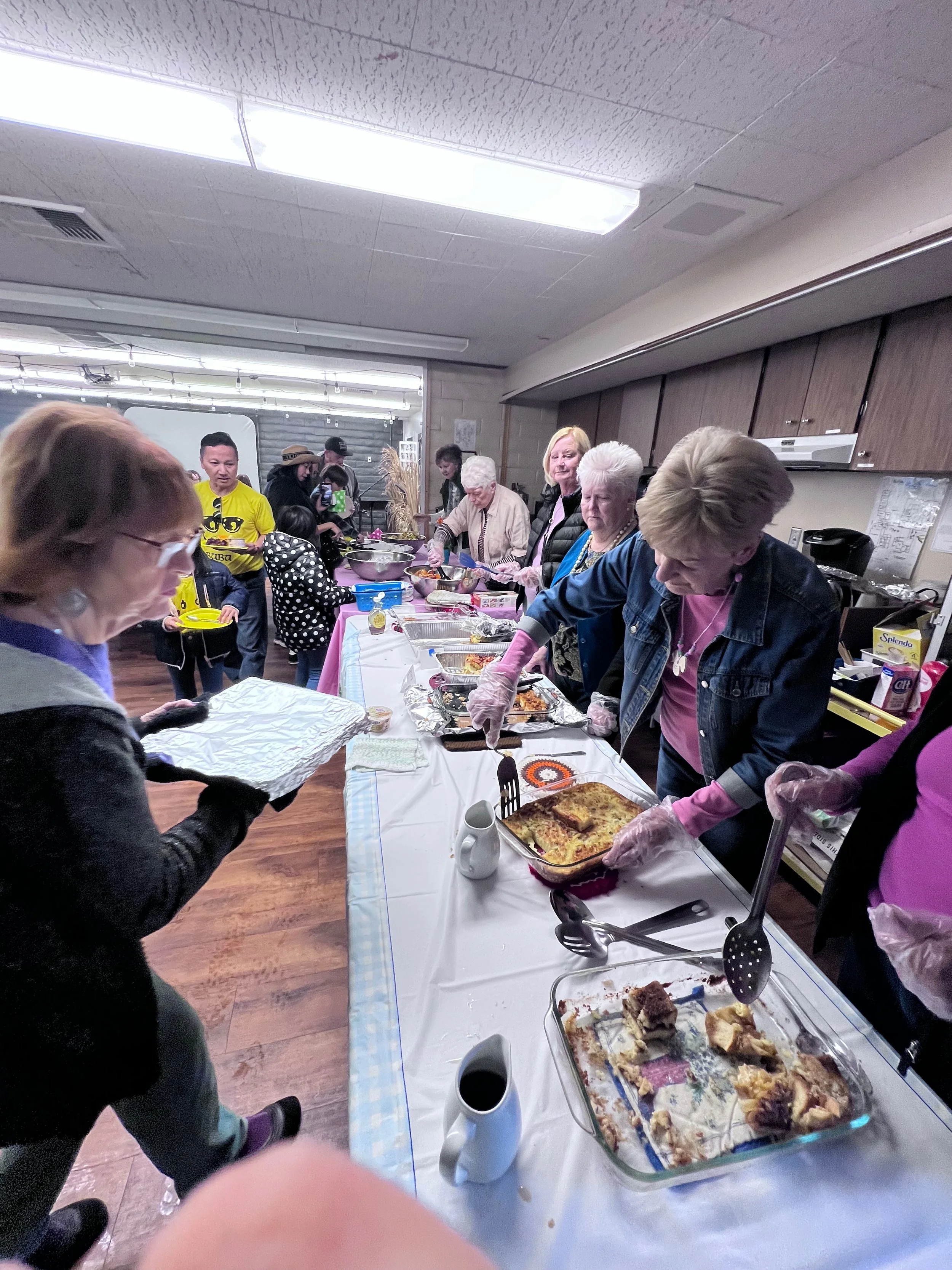 People serving and eating food at a communal gathering with a long table of various dishes and drinks.