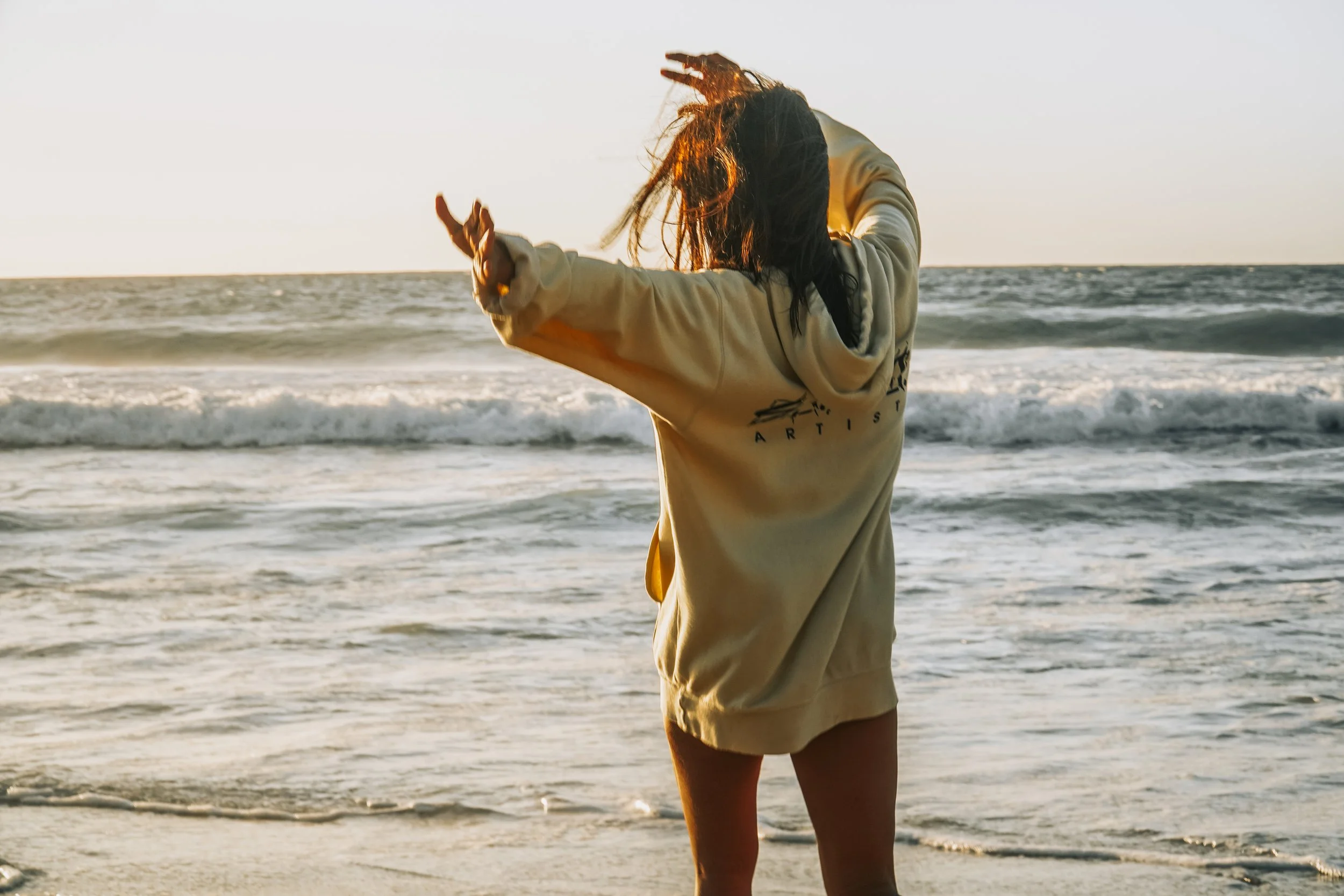 Person with dark hair dancing on the beach at sunset, facing the ocean with waves.