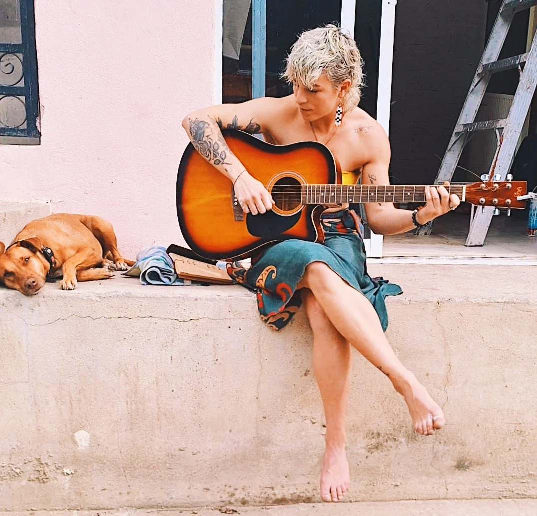 Woman playing guitar barefoot on storefront steps, with a pink background and a dog beside her