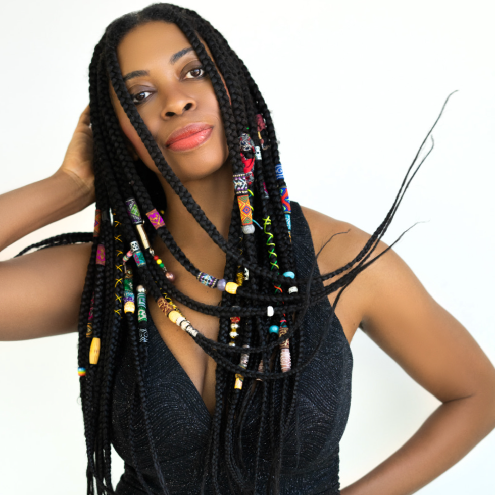 A woman with braided hair decorated with colorful beads, wearing a black sleeveless top, standing against a white background.