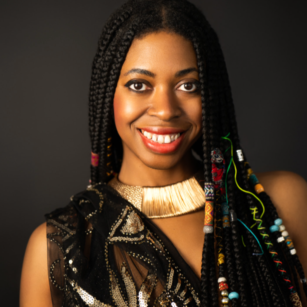 Portrait of a smiling woman with braided hair adorned with beads, wearing a gold choker necklace and a black embroidered dress against a dark background.