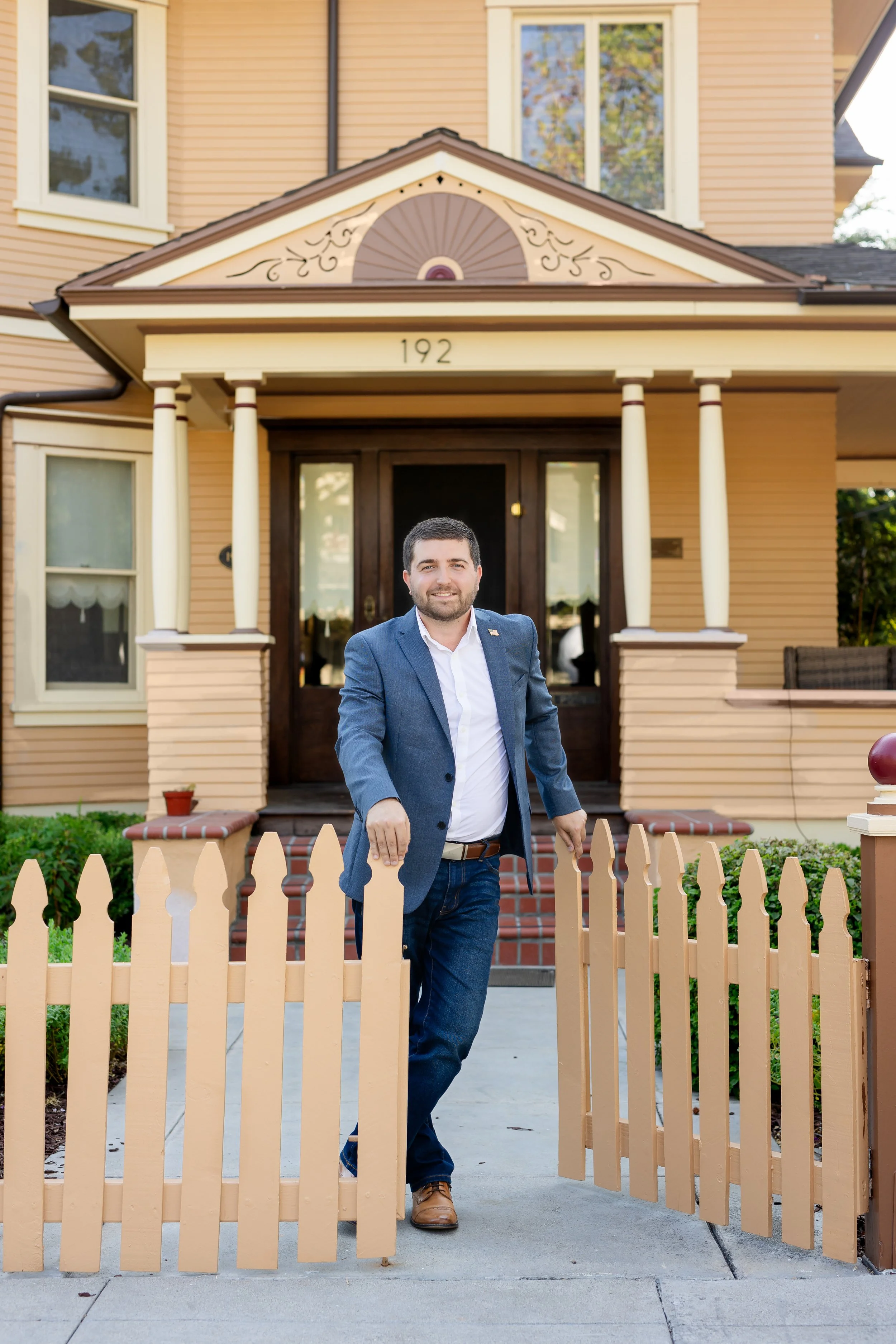 A man in a blue blazer and white shirt standing in a beige picket fence, smiling, in front of a yellow house with a porch and steps.