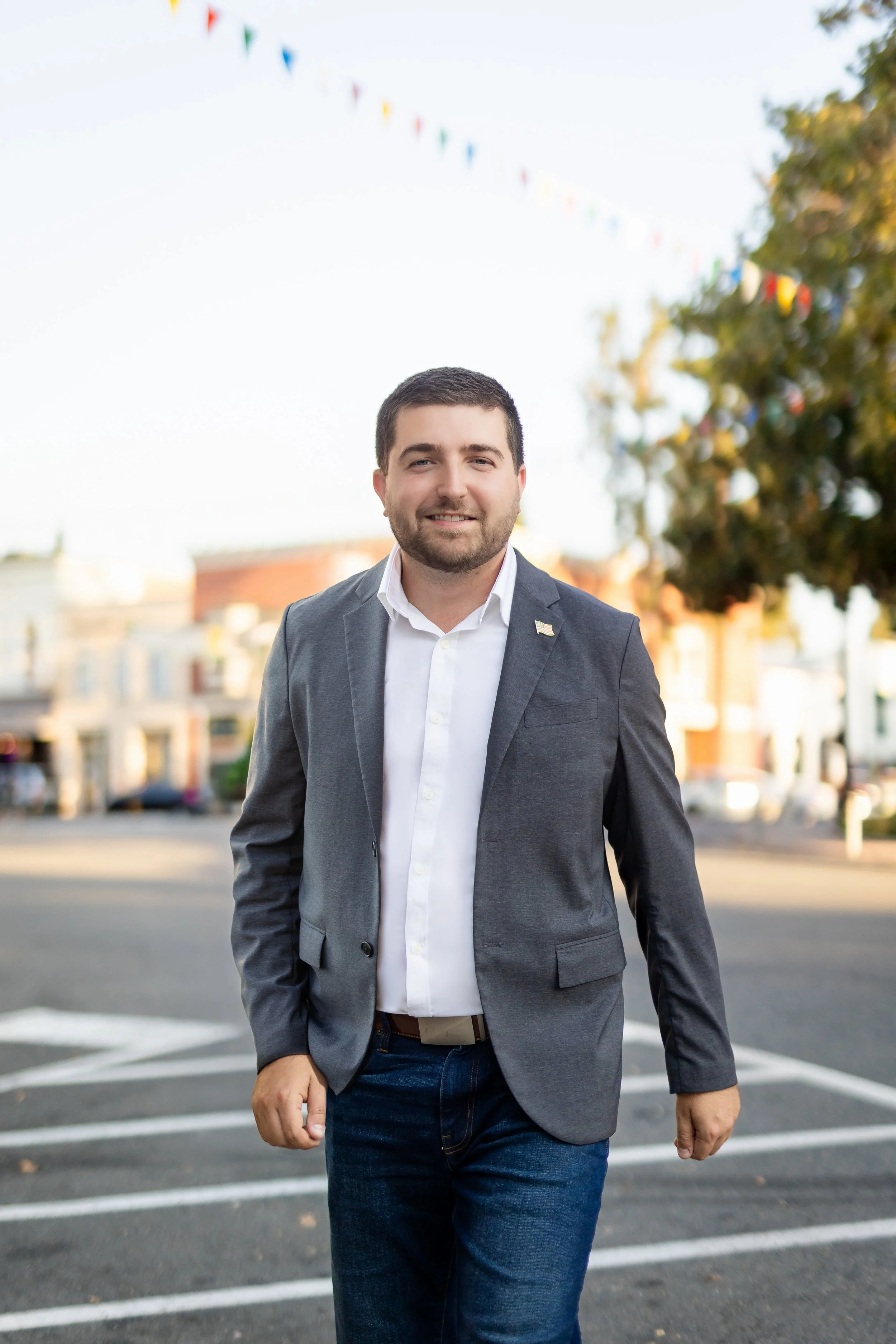 A man in a gray blazer and white shirt walking outdoors with a smile on his face.