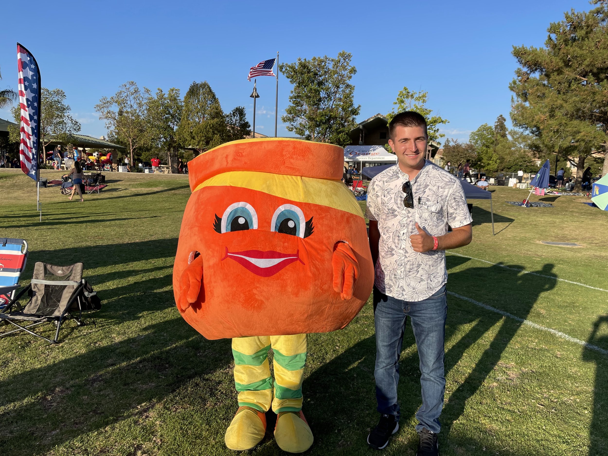 A man standing next to a person in a large, colorful costume of a smiling, anthropomorphic jar or container with big eyes and a wide smile, at an outdoor event on a sunny day.