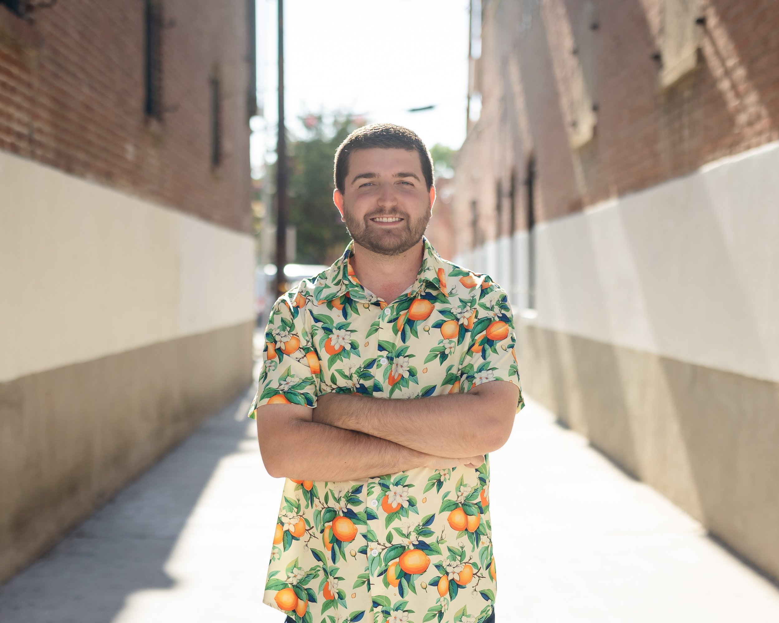 A man standing with arms crossed in an alleyway, smiling, wearing a colorful shirt with oranges and flowers, bright sunlight.