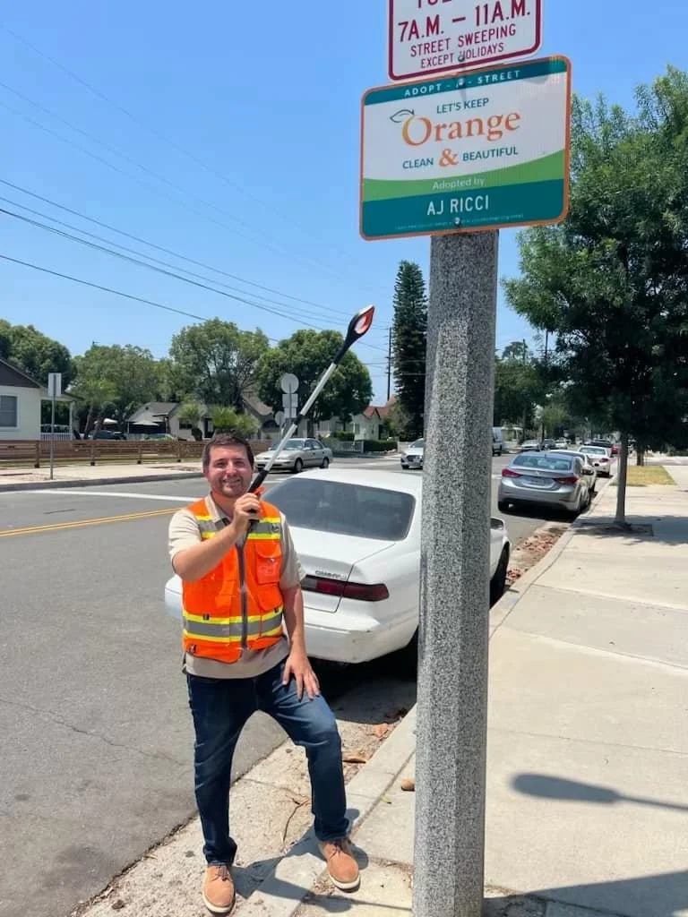 A man in an orange safety vest smiling and holding a trash picker, standing on a sidewalk next to a tree and a street sign in a suburban neighborhood.