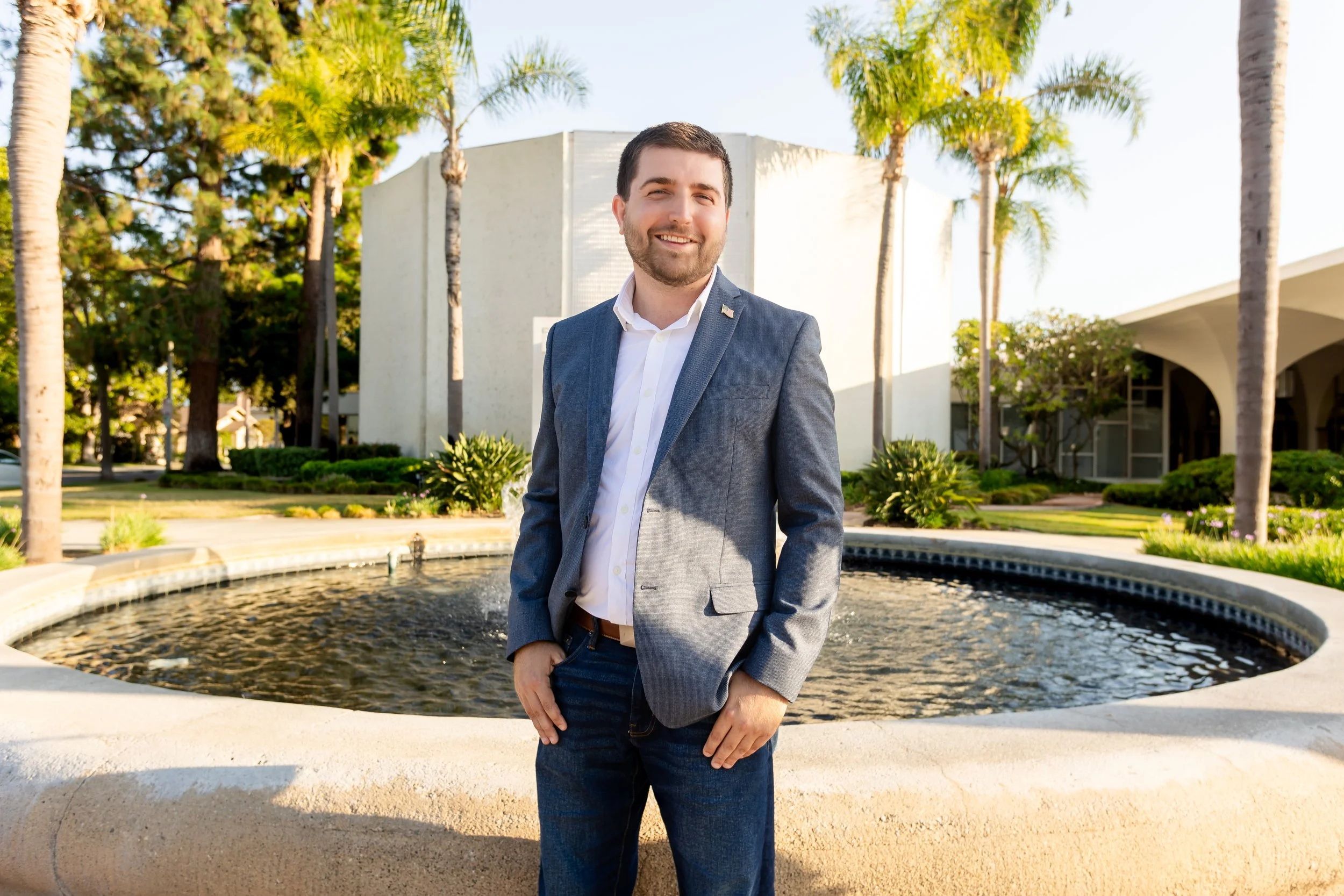 A man in a gray suit jacket and white shirt smiling and standing outdoors near a fountain with palm trees and buildings in the background.