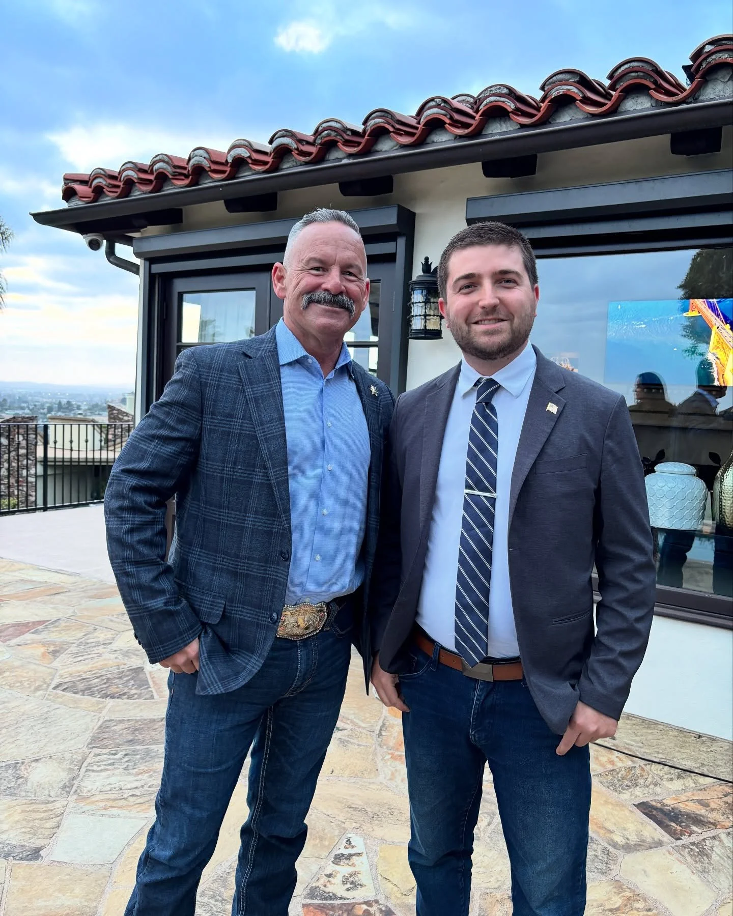 Two men in suits standing outdoors on a patio with city view in the background, smiling for the photo.