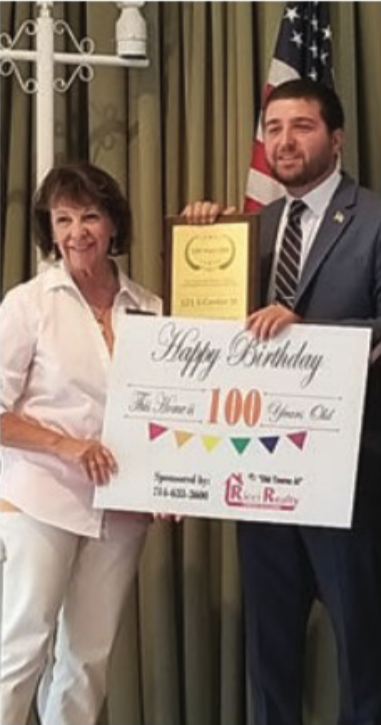 A woman and a man holding a large birthday sign and a certificate, standing in front of curtains with an American flag in the background.
