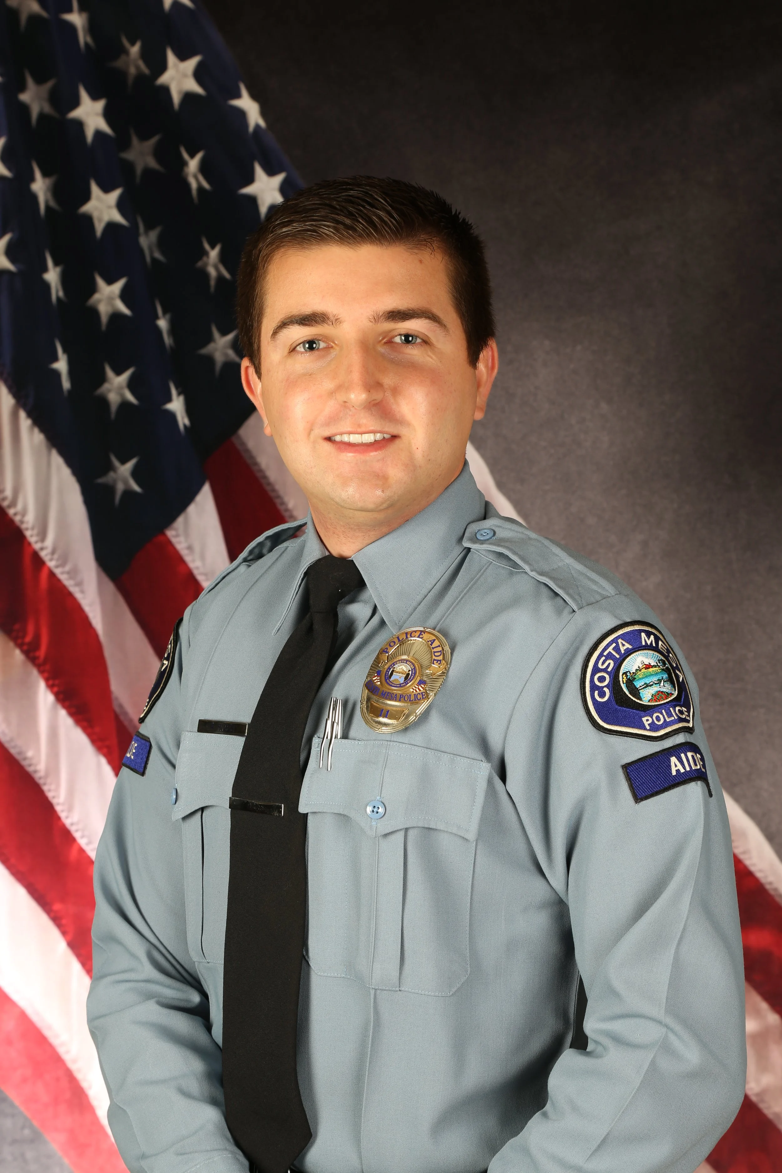 officer in uniform posing in front of an American flag. The officer is smiling and has a badge and patches on his uniform, which includes a shoulder patch reading "Costa Mesa Police" and an "Aide" patch. The background is dark grey.