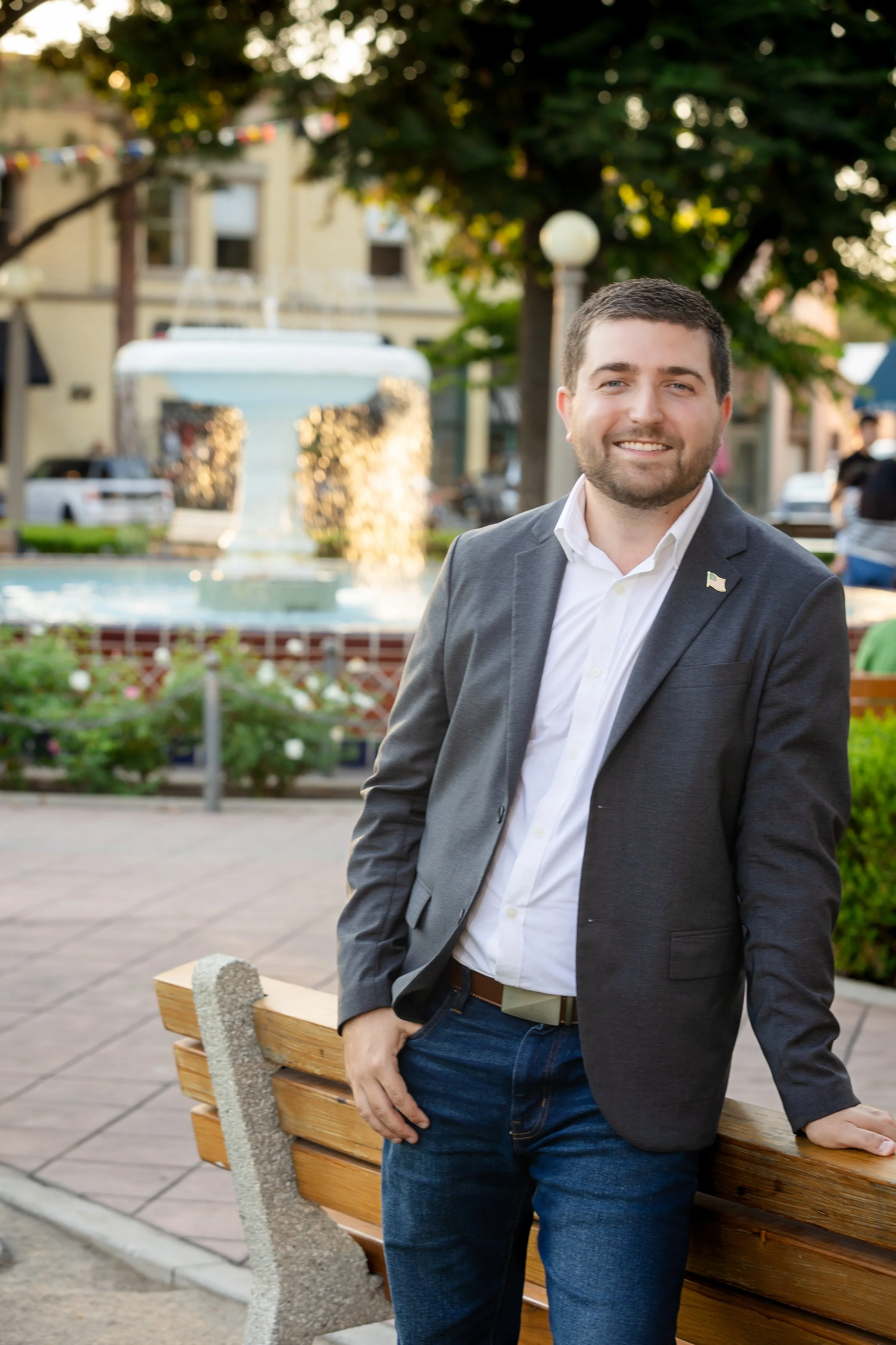 A man in a gray blazer and white shirt standing outside near a wooden park bench with a fountain and trees in the background.