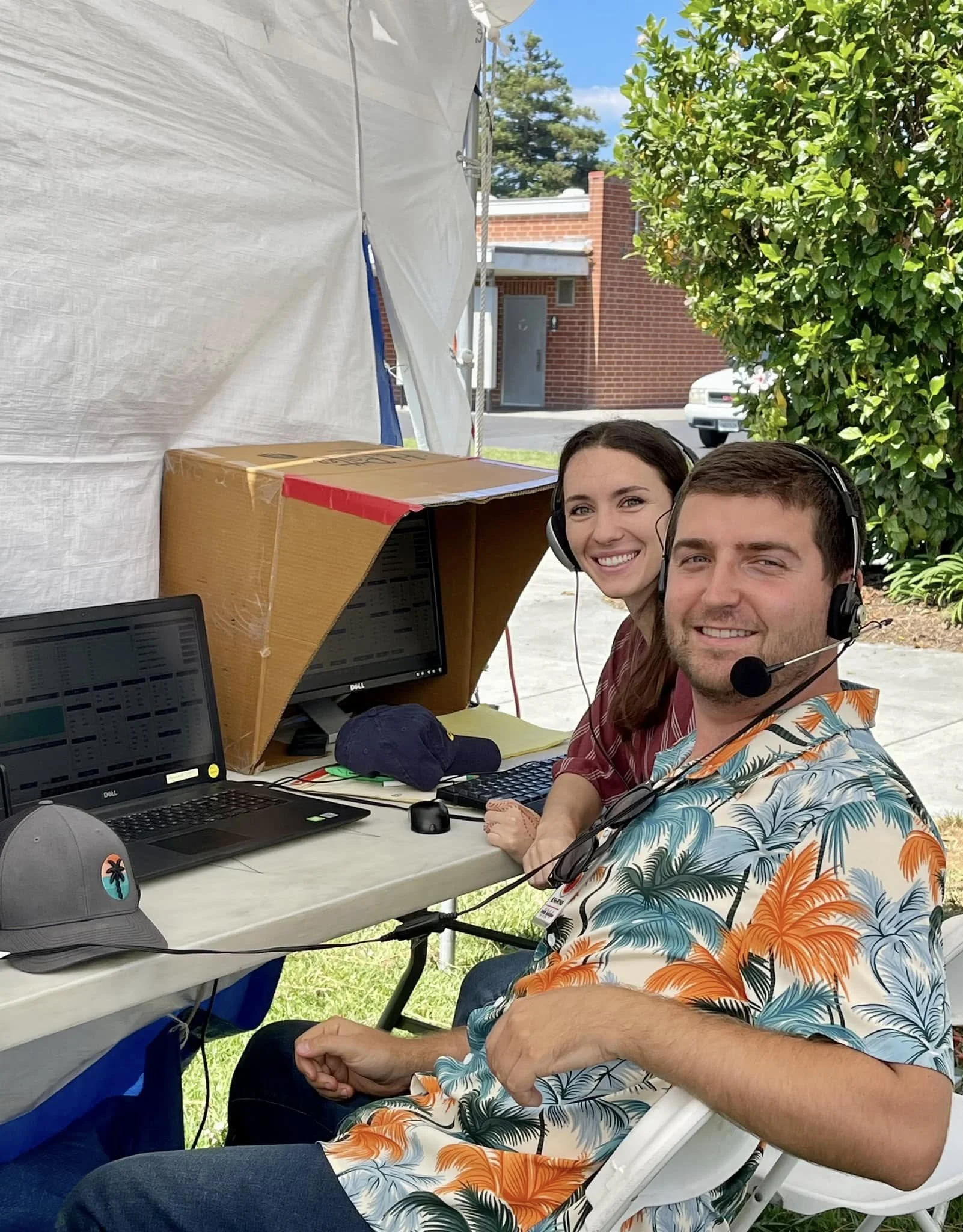 Two smiling call center workers with headsets working at a table inside a tent outdoors, with laptops, monitors, and a cardboard privacy screen.