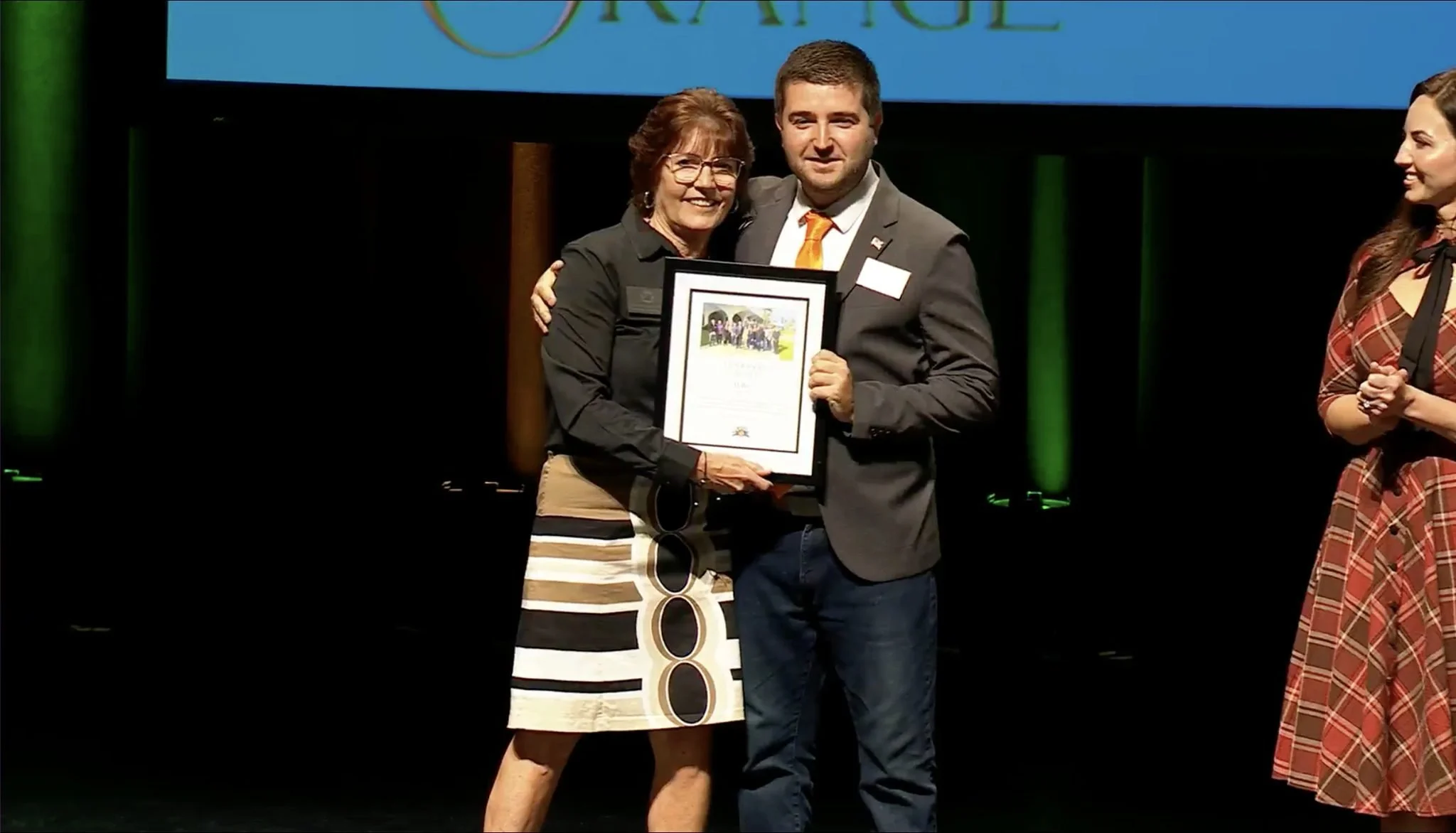 A woman and a man holding an award certificate on stage at an event, with a woman in a brown plaid dress standing beside them and smiling.