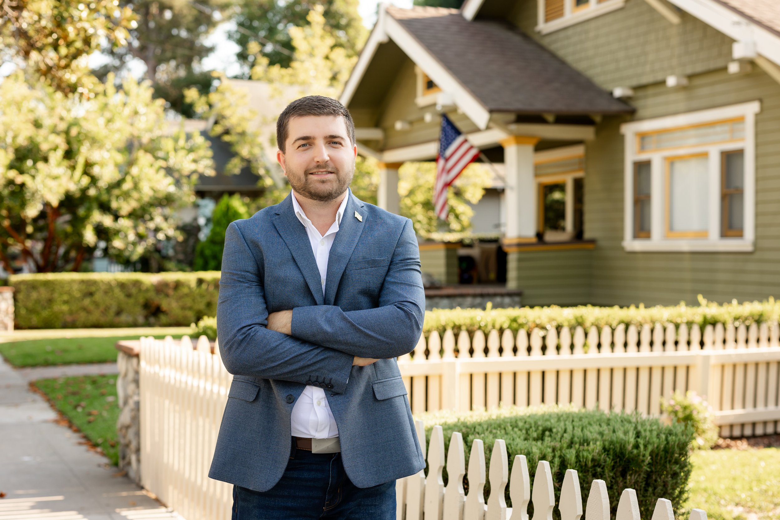 A man in a blue blazer and white shirt standing with arms crossed in front of a house with an American flag in the yard.