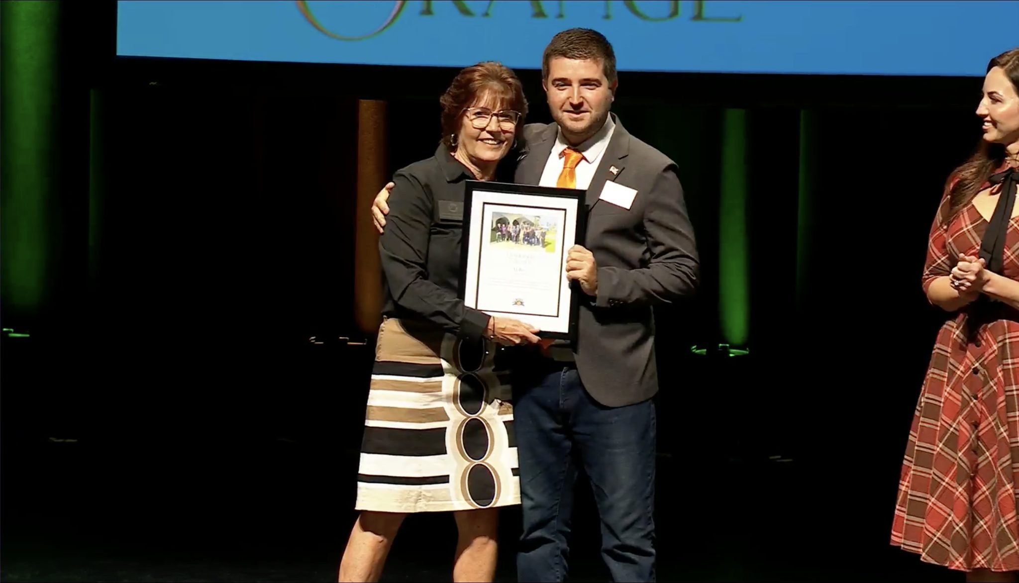 Woman in black jacket receiving award from a man in gray suit on stage, two other women on stage, background dark with green lighting.