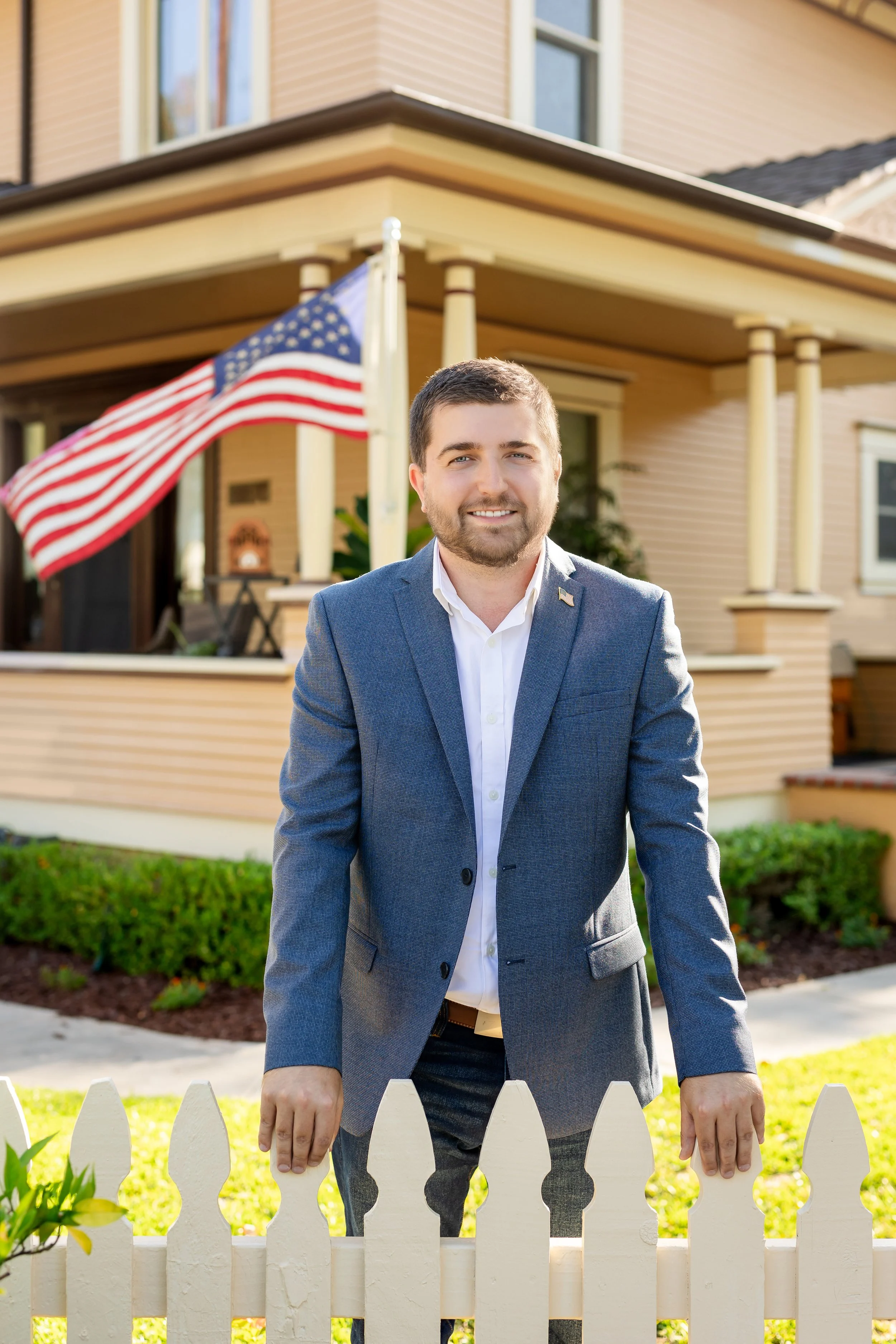 A man in a blue suit and white shirt standing behind a white picket fence in front of a beige house with an American flag on the porch.