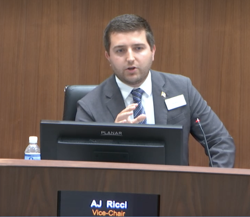 A man in a suit speaking at a conference or panel, seated behind a desk with a computer monitor and a water bottle, with a nameplate reading 'AJ Ricci Vice-Chair'.