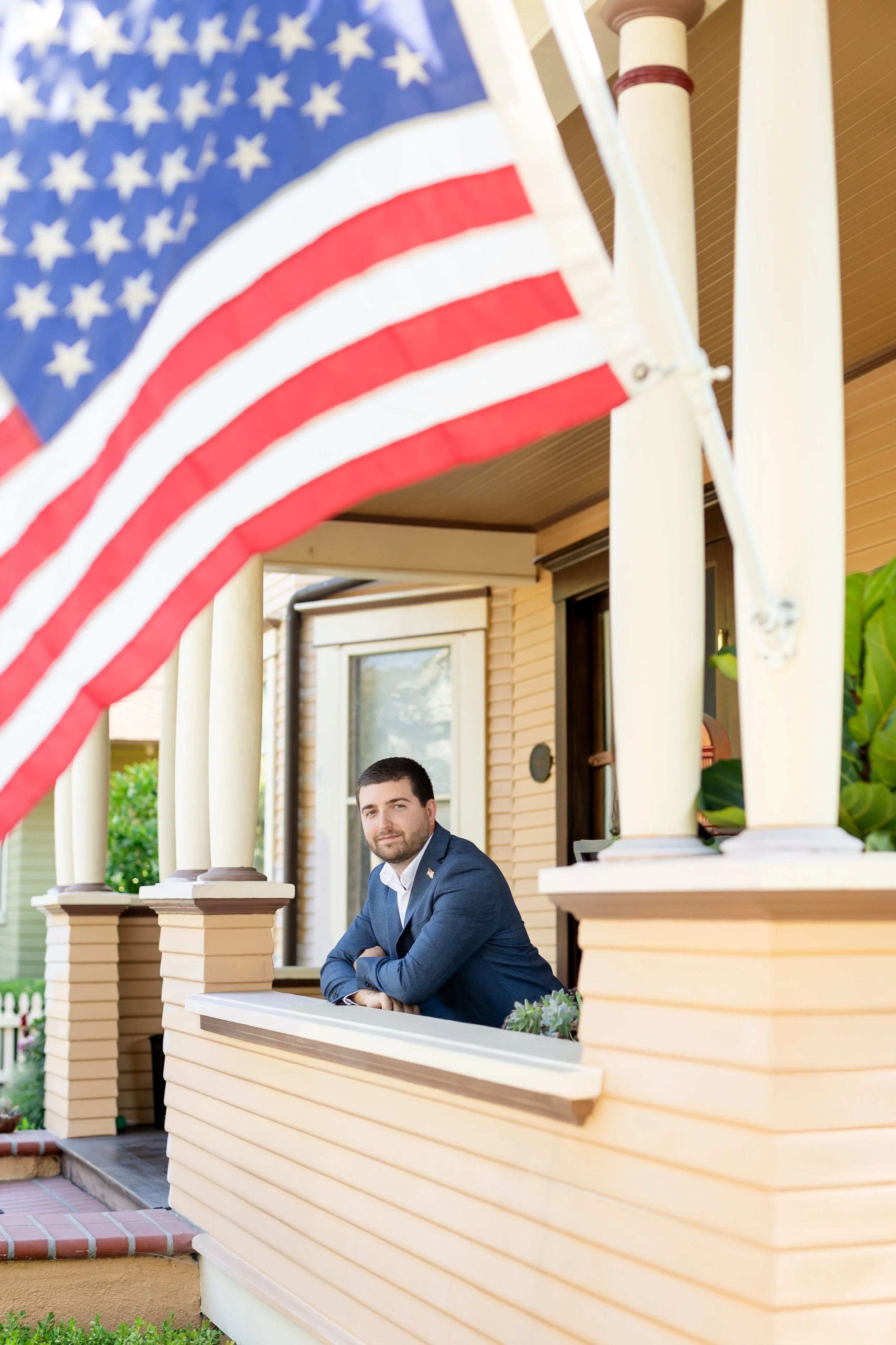 A man in a navy blazer leaning on a porch railing with an American flag in the foreground.