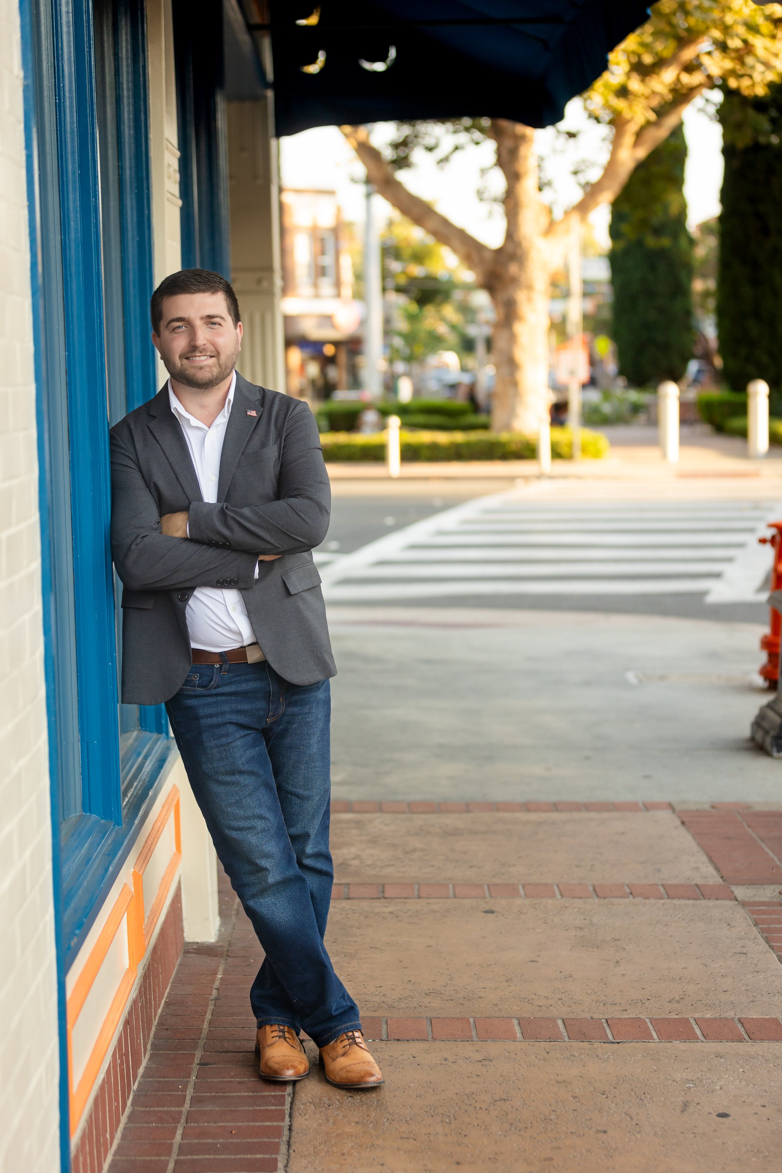 A man in a gray blazer and jeans standing with arms crossed, leaning against a building on a sidewalk in a city street.