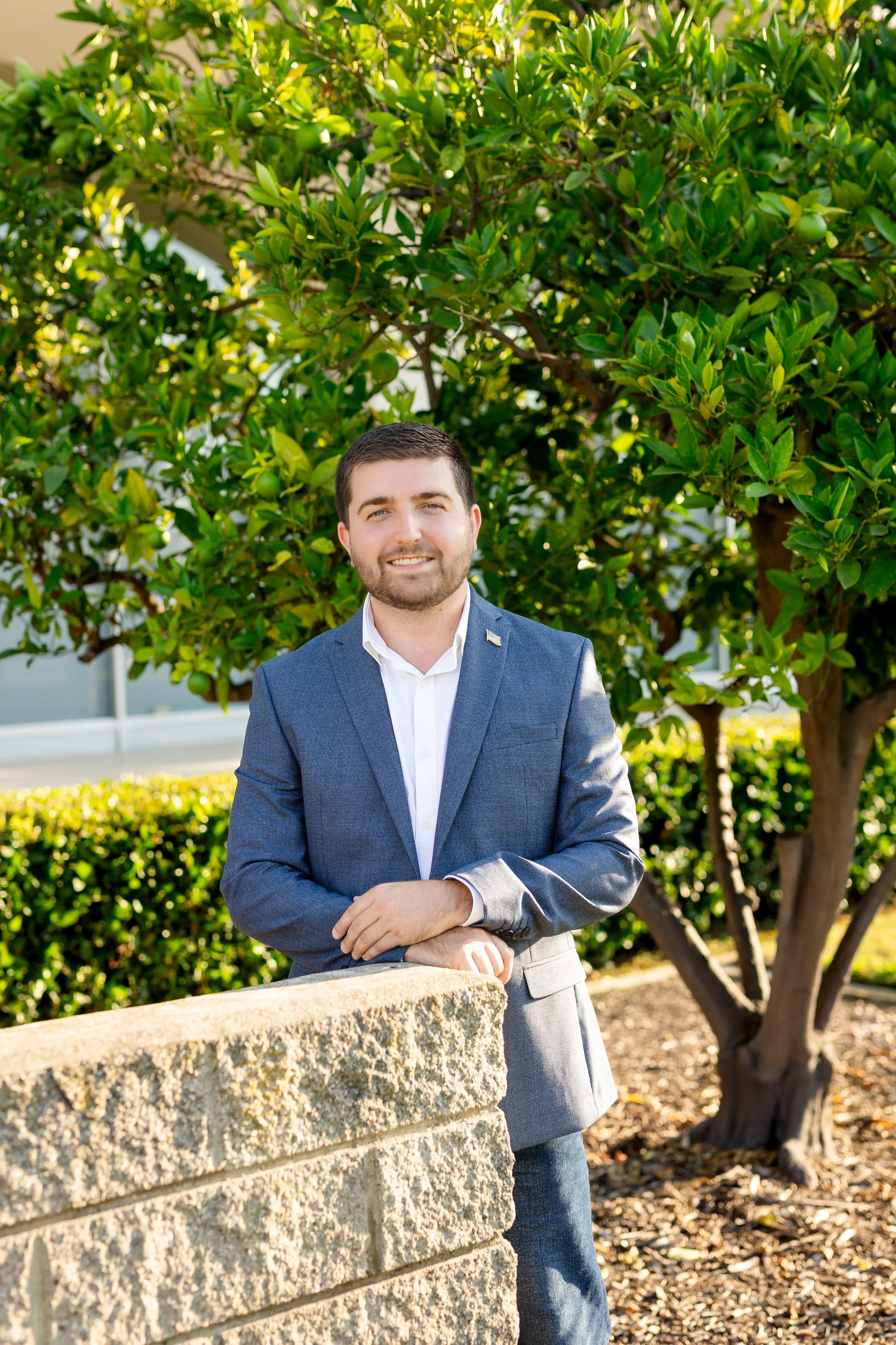 A man in a blue blazer and white shirt standing outdoors, leaning on a brick wall with a large green tree behind him.