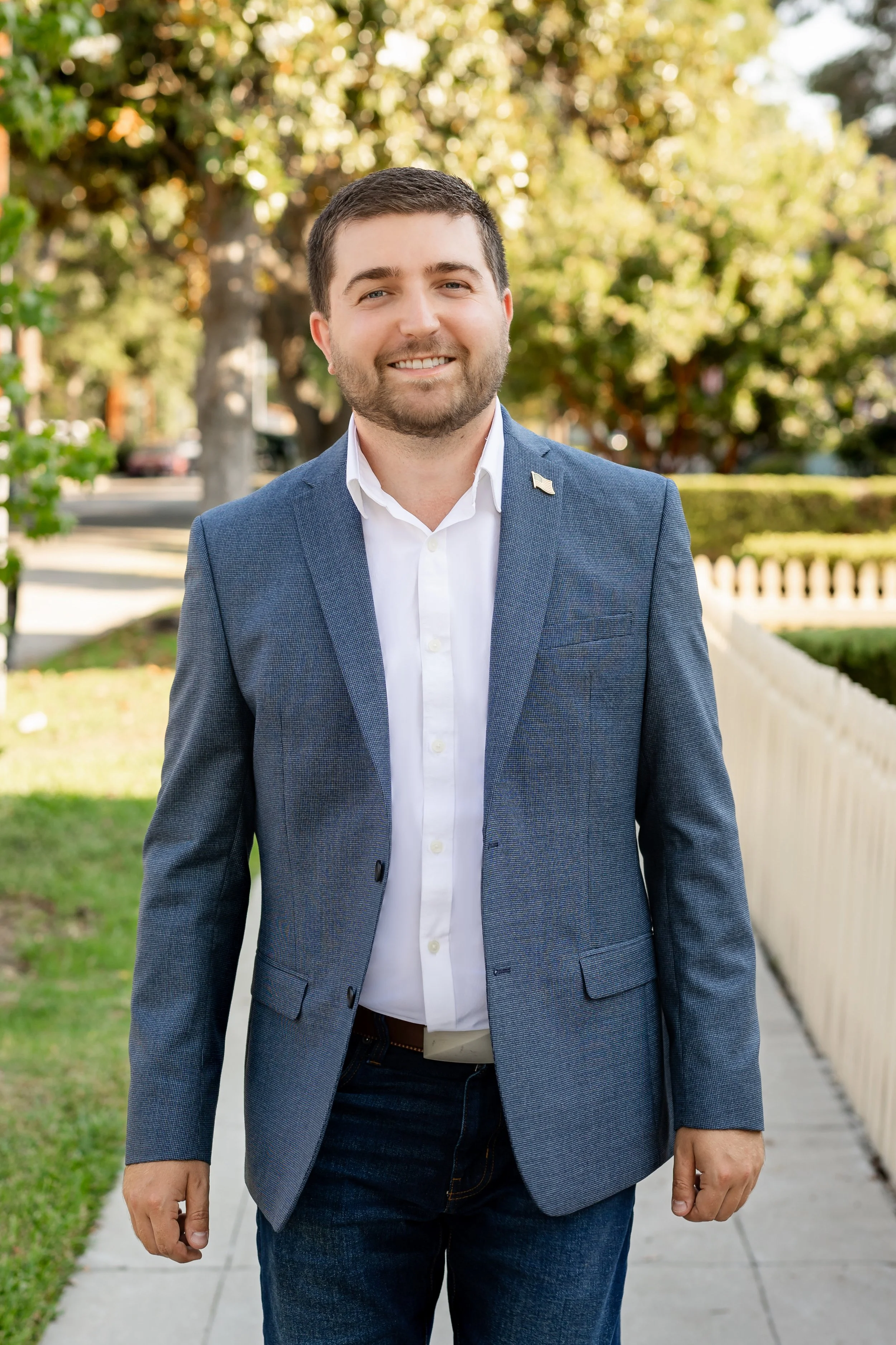 A man in a blue blazer and white shirt walking outdoors on a sidewalk with a white picket fence and green trees in the background.