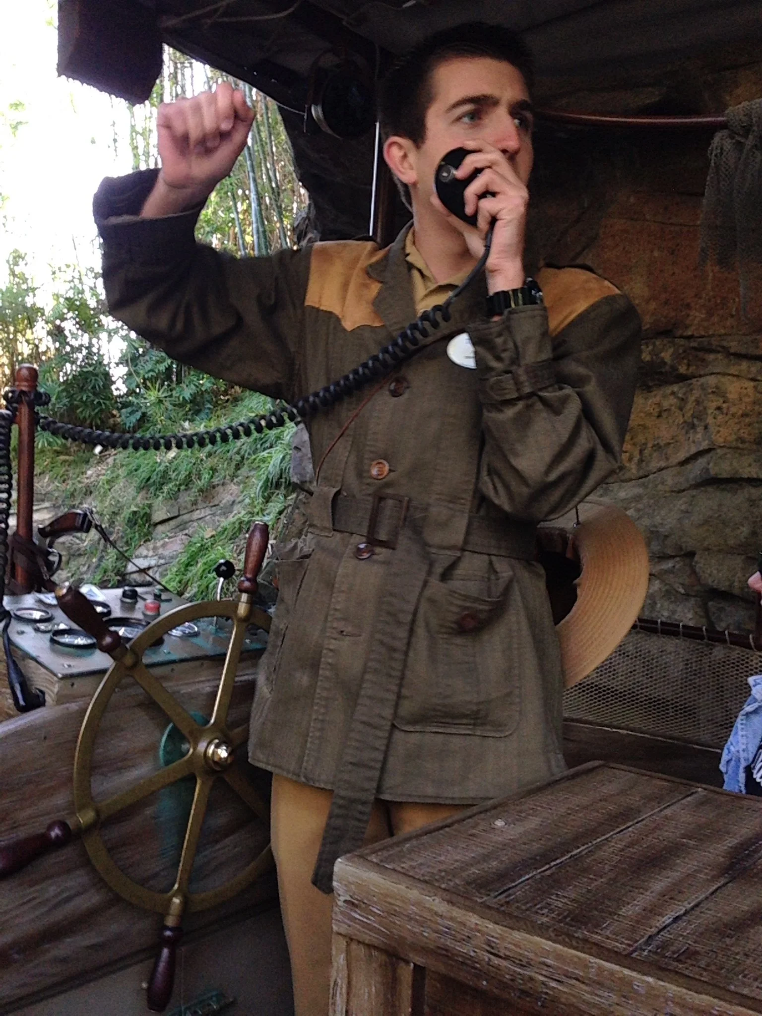 A person dressed in a vintage pilot or explorer costume holding a telephone receiver to their mouth, standing in a rustic setting with a ship's wheel and navigation equipment nearby.