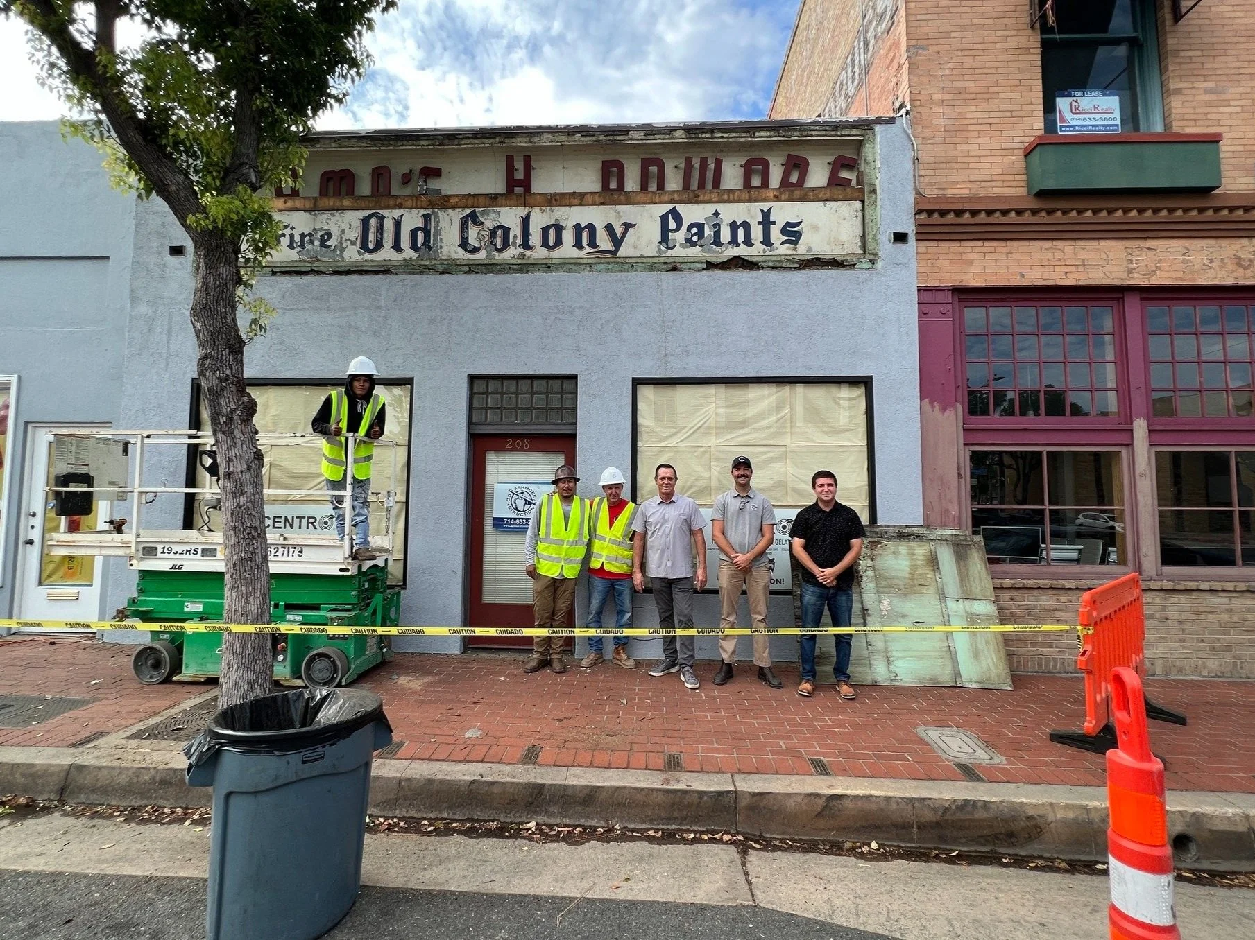 Group of five men standing outside a building with a sign that reads 'Old Colony Paints', some wearing safety vests and hard hats, with construction equipment and caution tape in front of them.