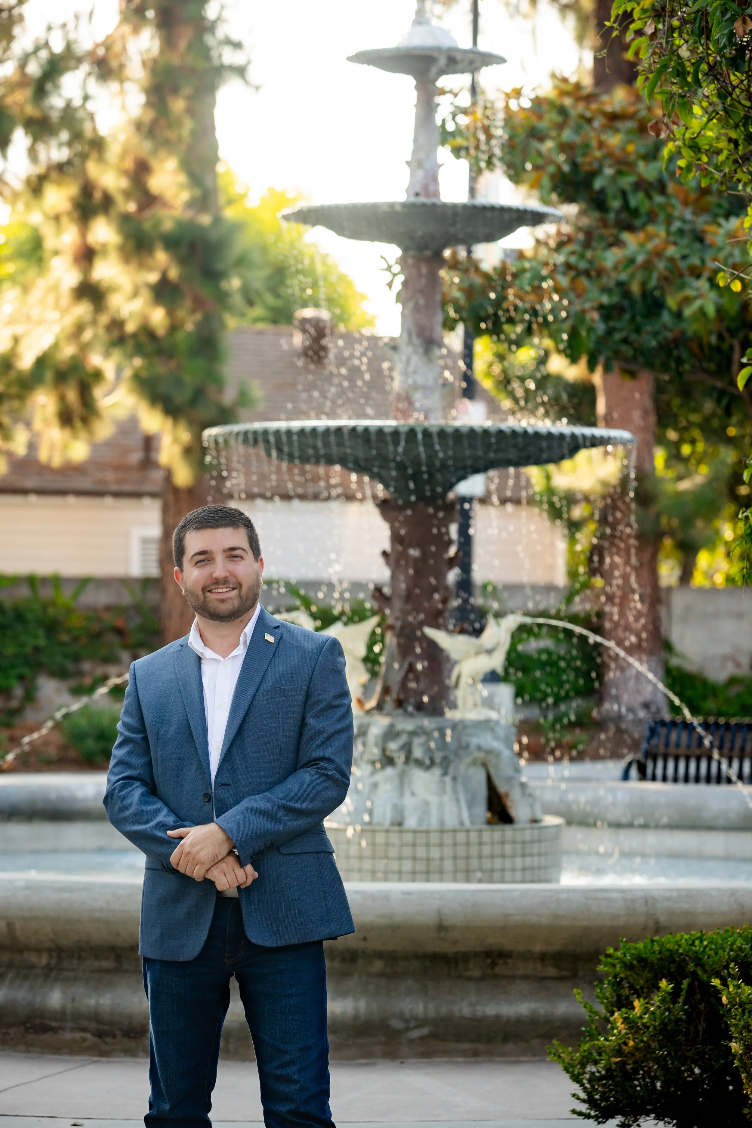 A man in a blue blazer and jeans standing in front of a multi-tiered fountain in a park, with greenery and trees in the background.