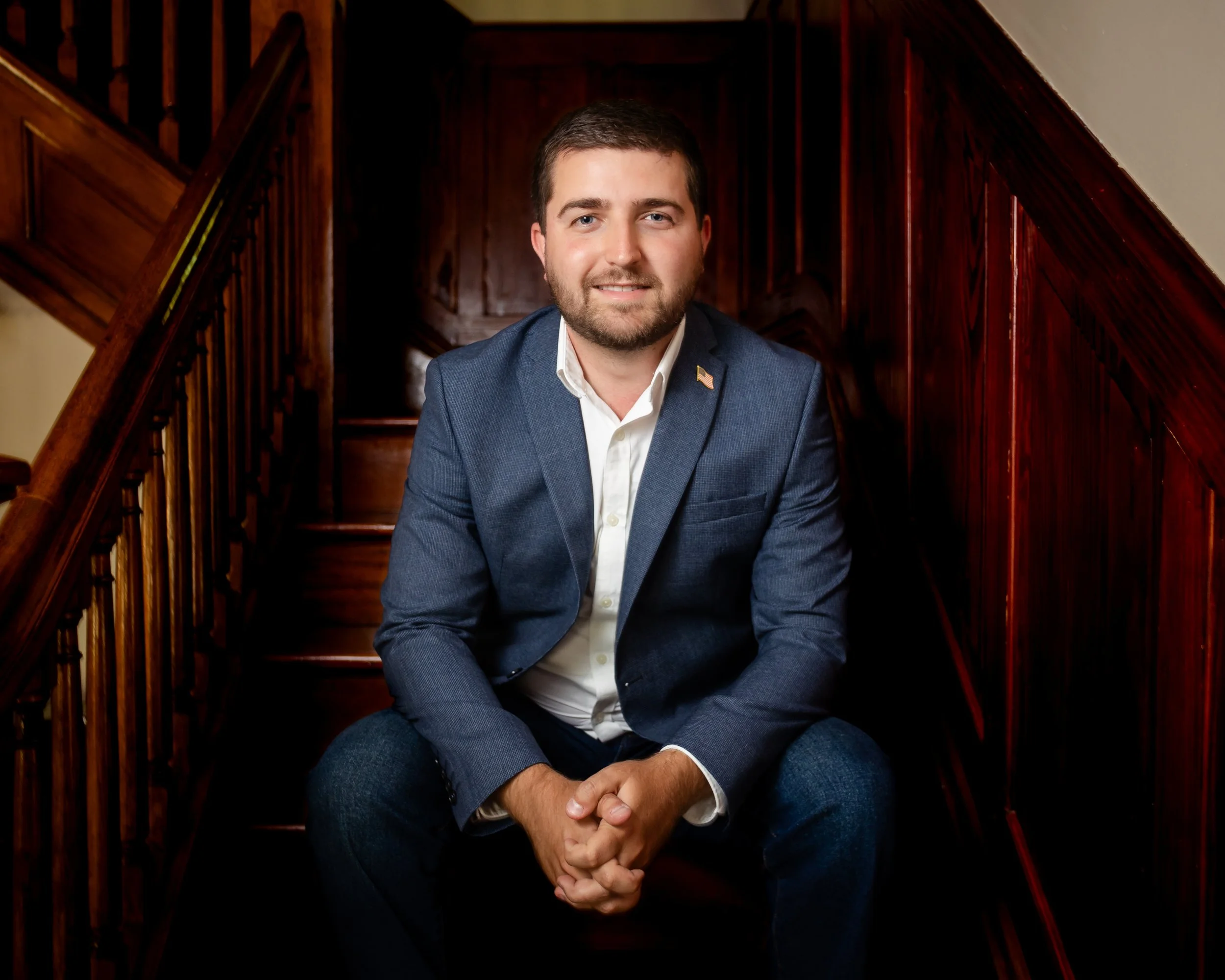 A man with a beard and short hair, wearing a navy blazer and white shirt, sitting on wooden stairs inside a building.