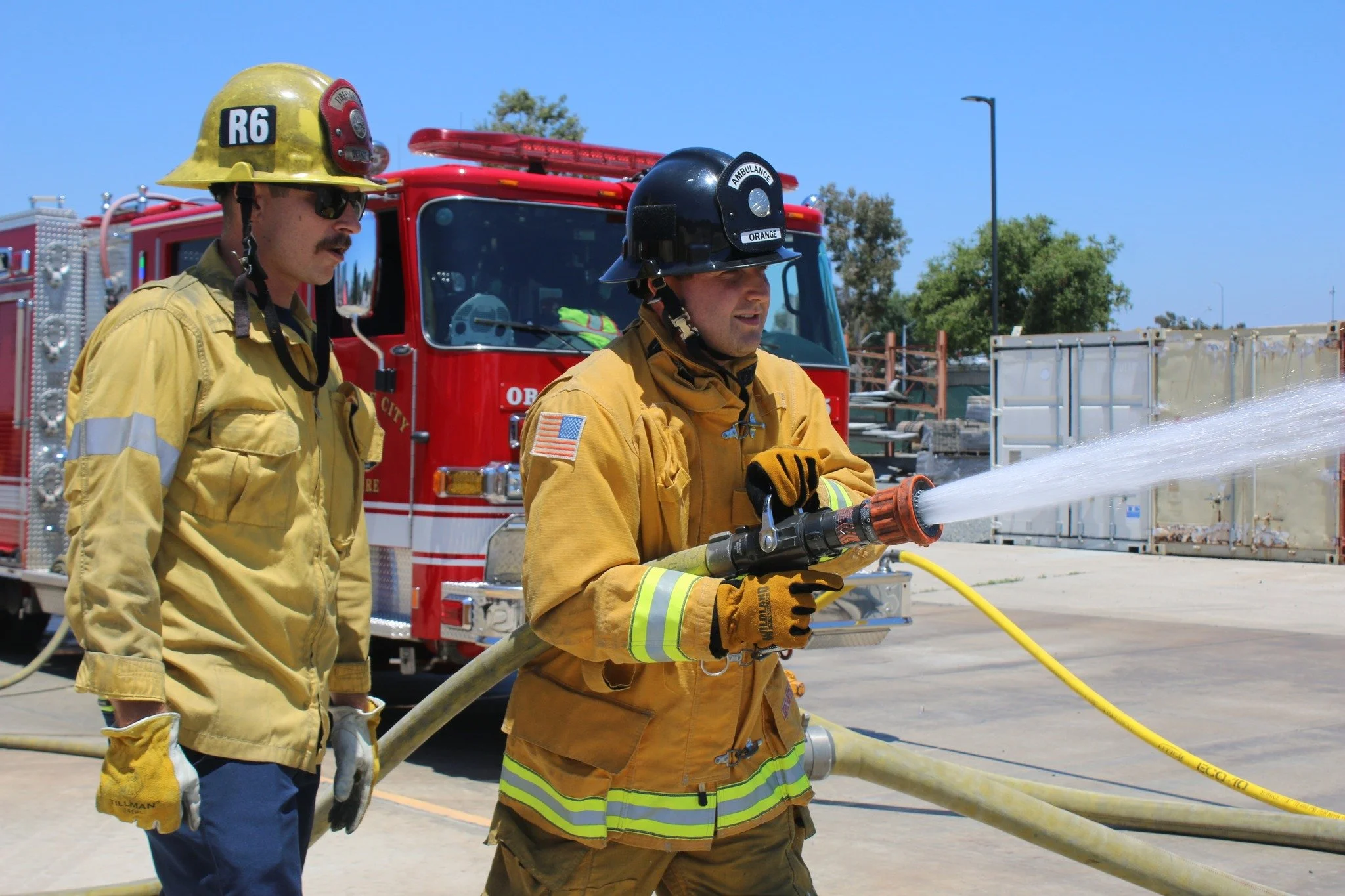 Two firefighters: one in yellow gear, and one actively spraying water, with a red fire truck in the background on a sunny day.