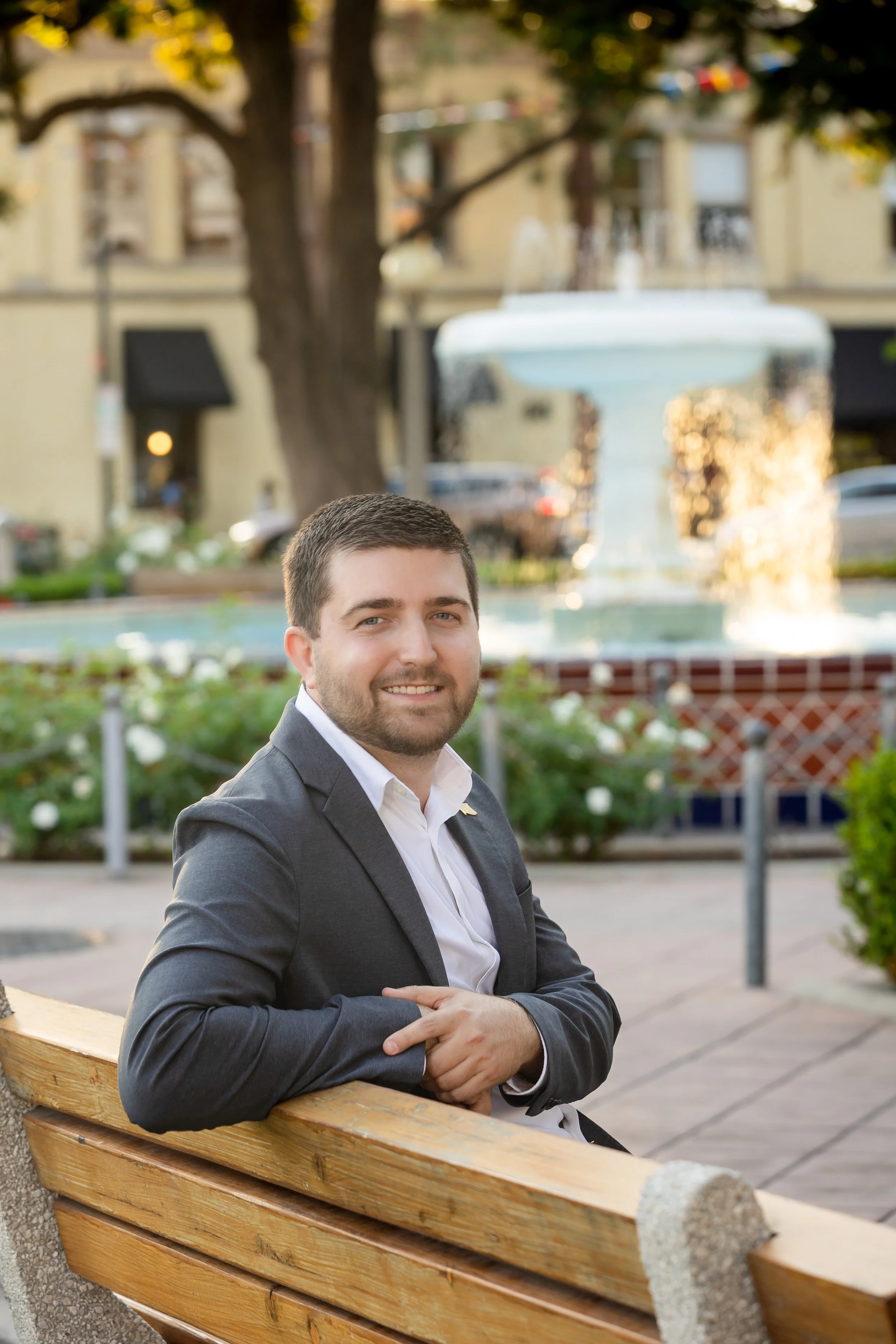 A man with short dark hair and a beard sitting on a wooden park bench in front of a fountain, smiling at the camera, dressed in a gray suit jacket and white shirt, in an outdoor urban setting.