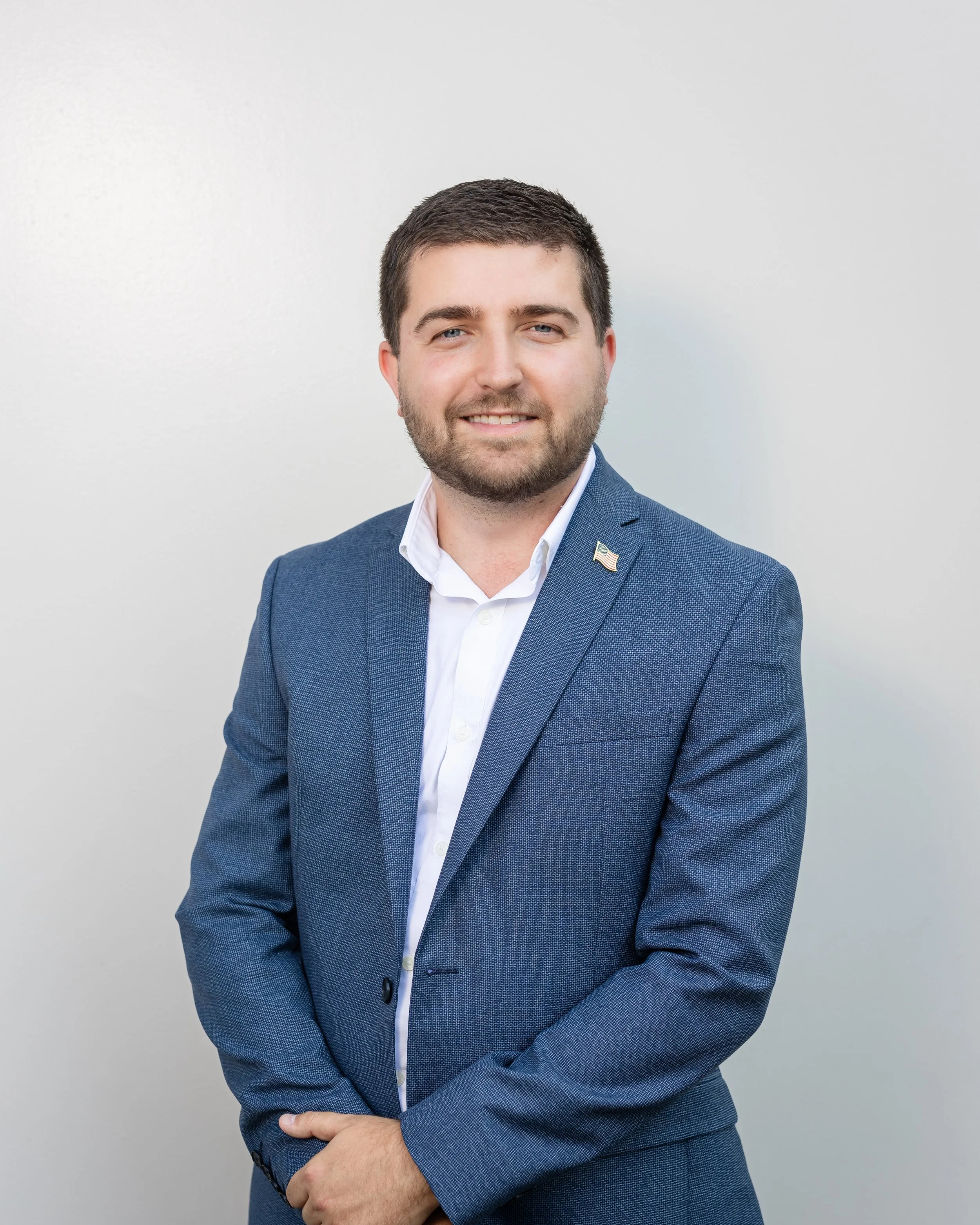 A man with short brown hair, beard, and light skin, dressed in a white shirt and blue suit, smiling at the camera with his hands clasped in front of him, standing against a plain white background.