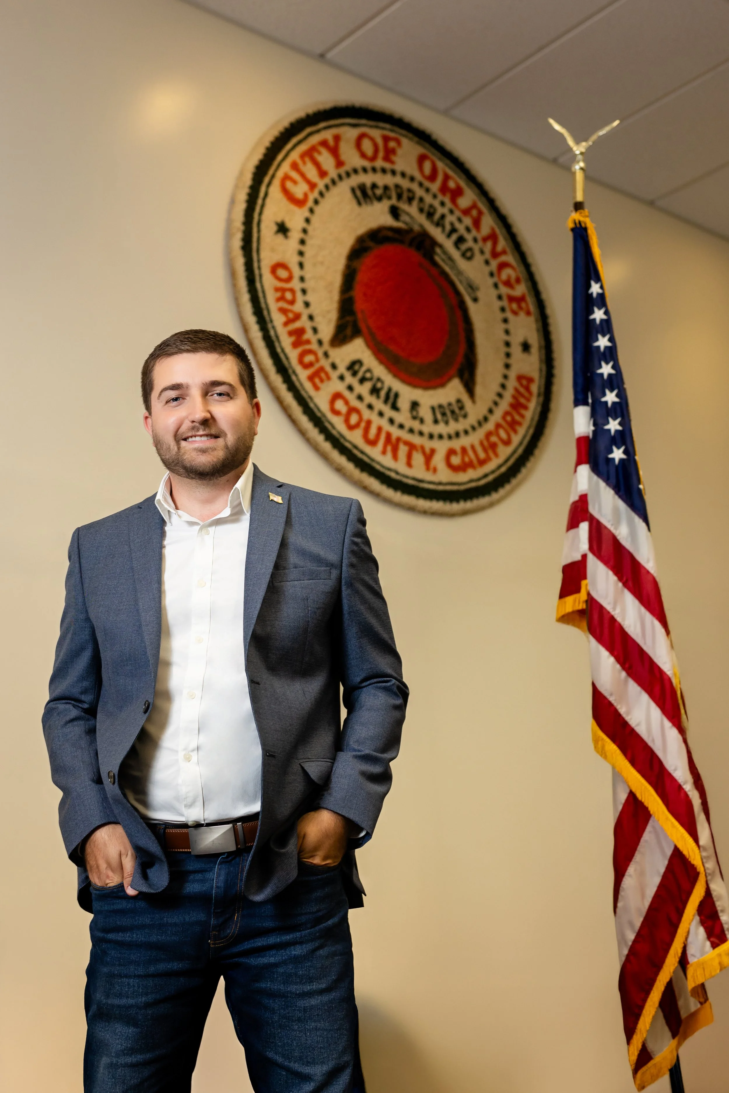 A man in a suit standing next to an American flag in front of a city of Orange, California sign with a lemon logo.