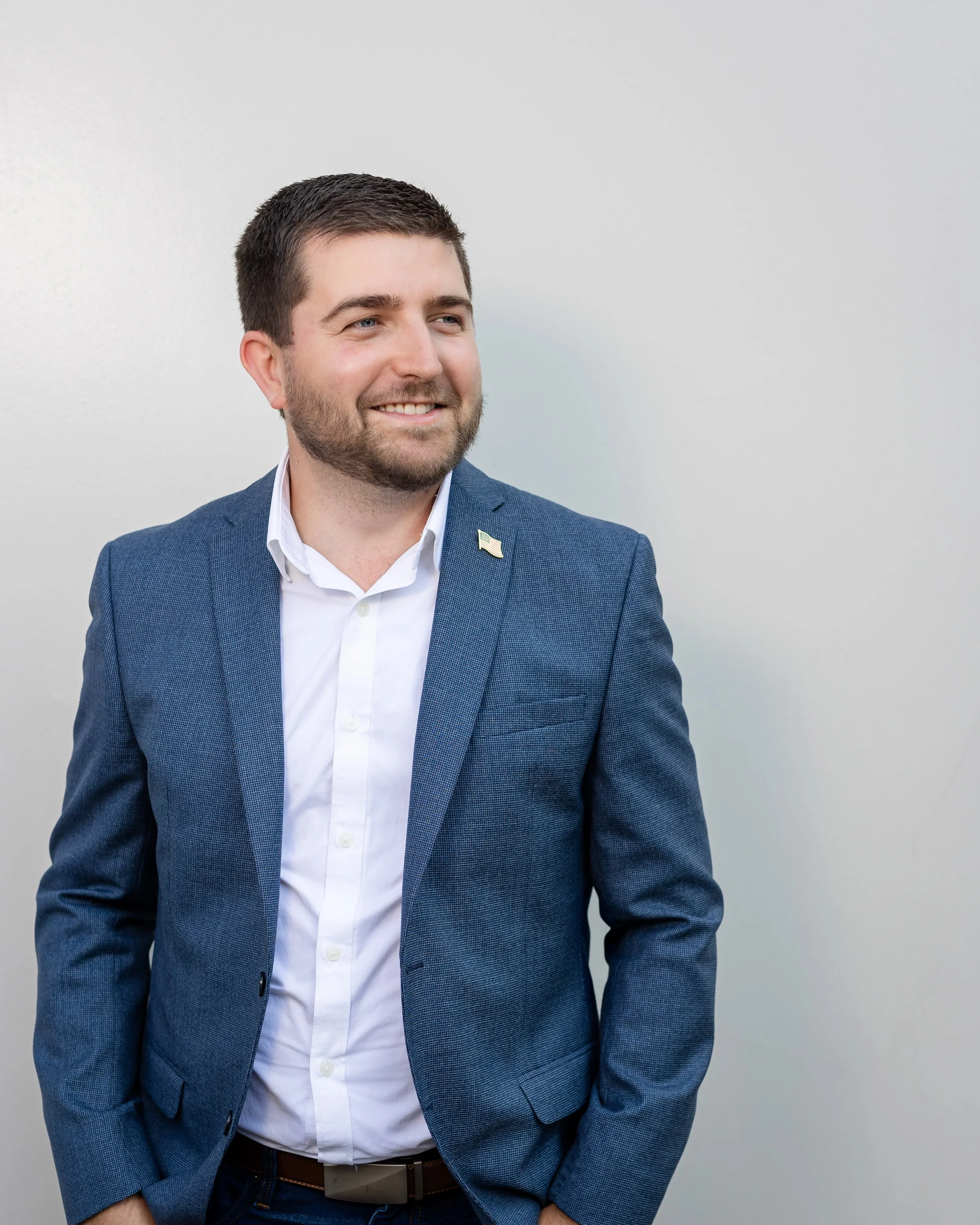 Man in a blue suit and white shirt smiling, standing against a plain white wall.