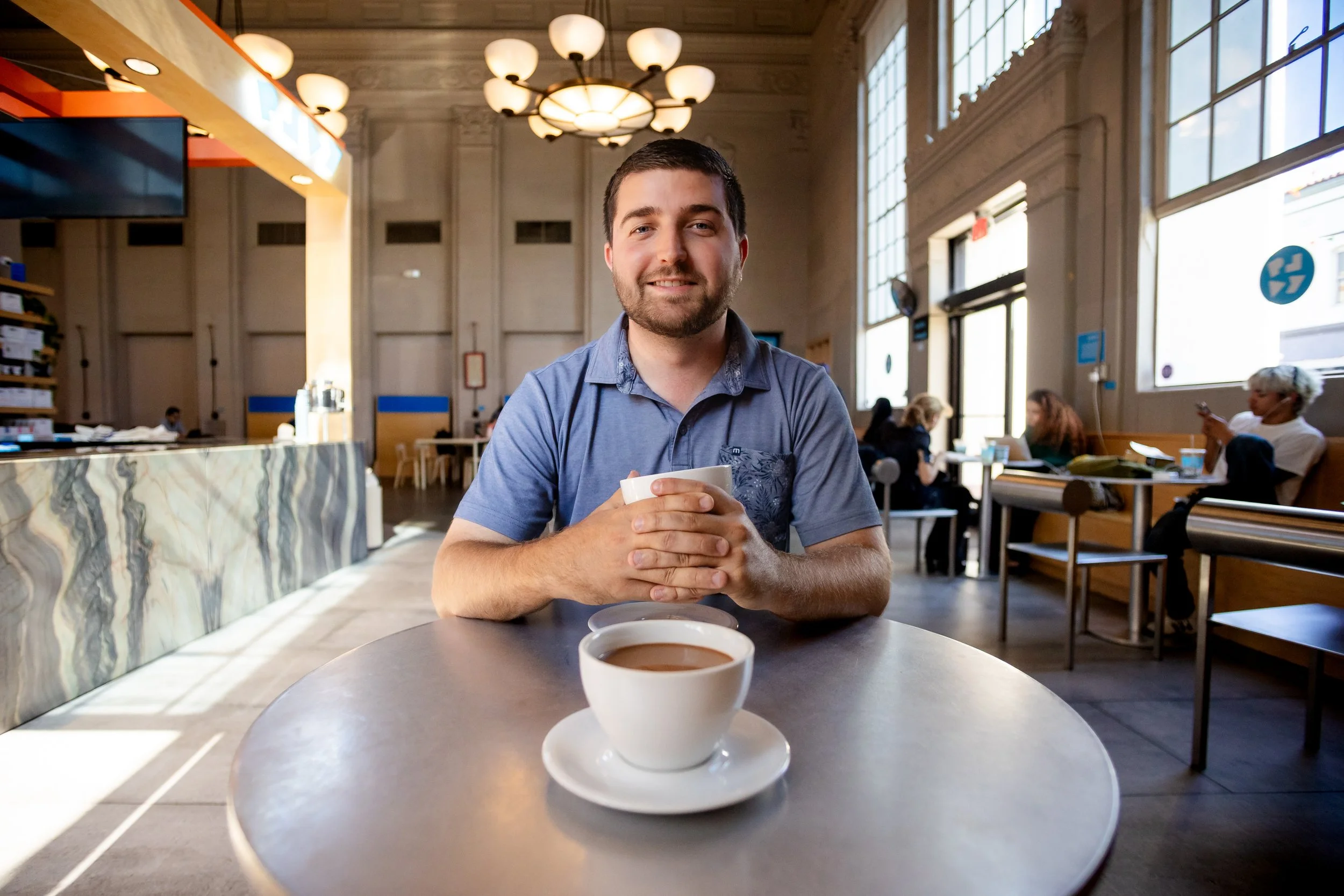 A man with short dark hair and a beard, wearing a blue shirt, sitting at a round table with two cups of coffee in front of him, smiling at the camera in a bright, spacious café with large windows and other patrons in the background.