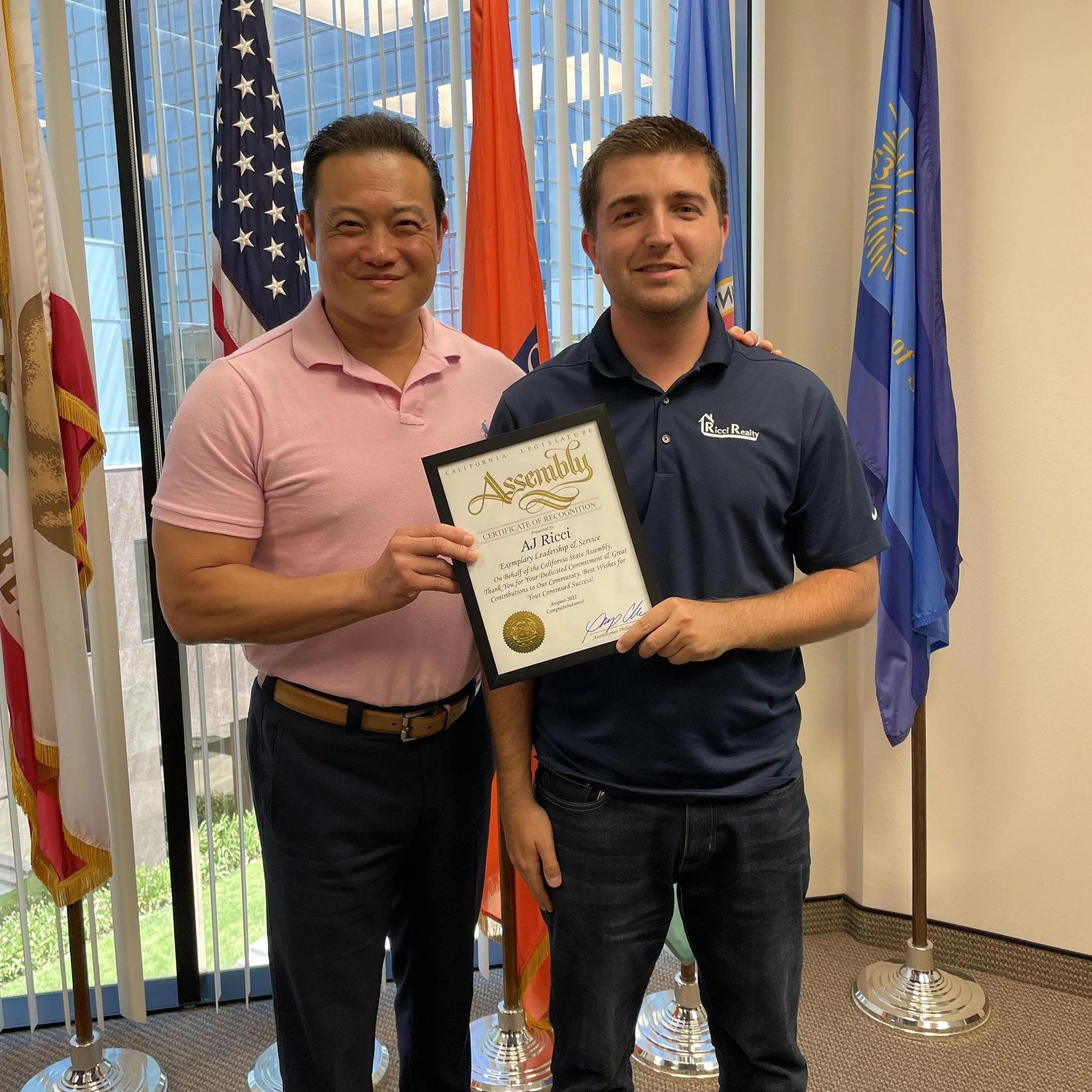 Two men standing inside, one holding a framed certificate. Flags are visible in the background.
