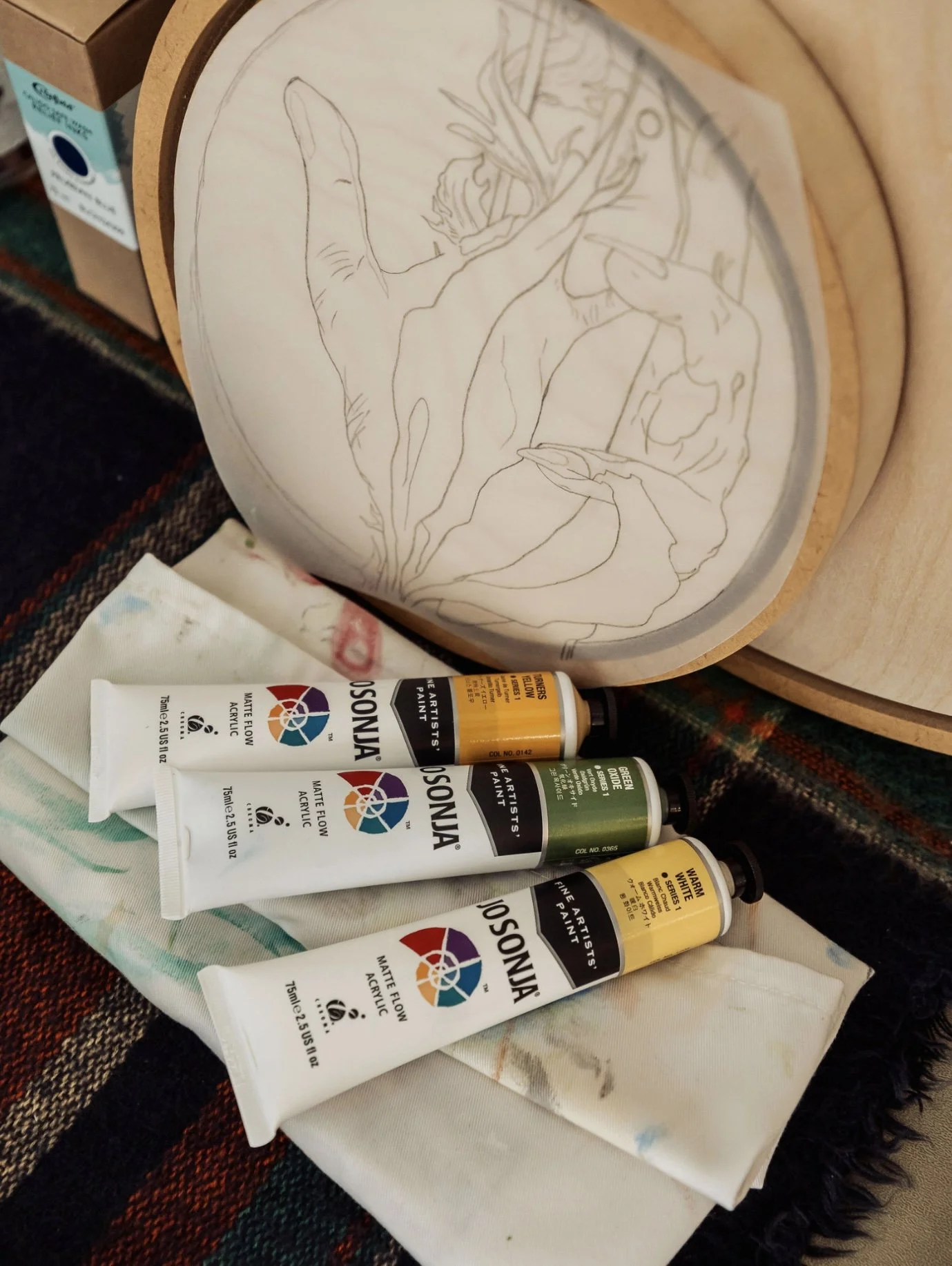 Paint tubes, including yellow, green, and white, are placed on a cloth on a table next to an unfinished wooden art piece with a sketch of an abstract clawed hand on tracing paper. There's a cardboard box and a dark woven fabric in the background.