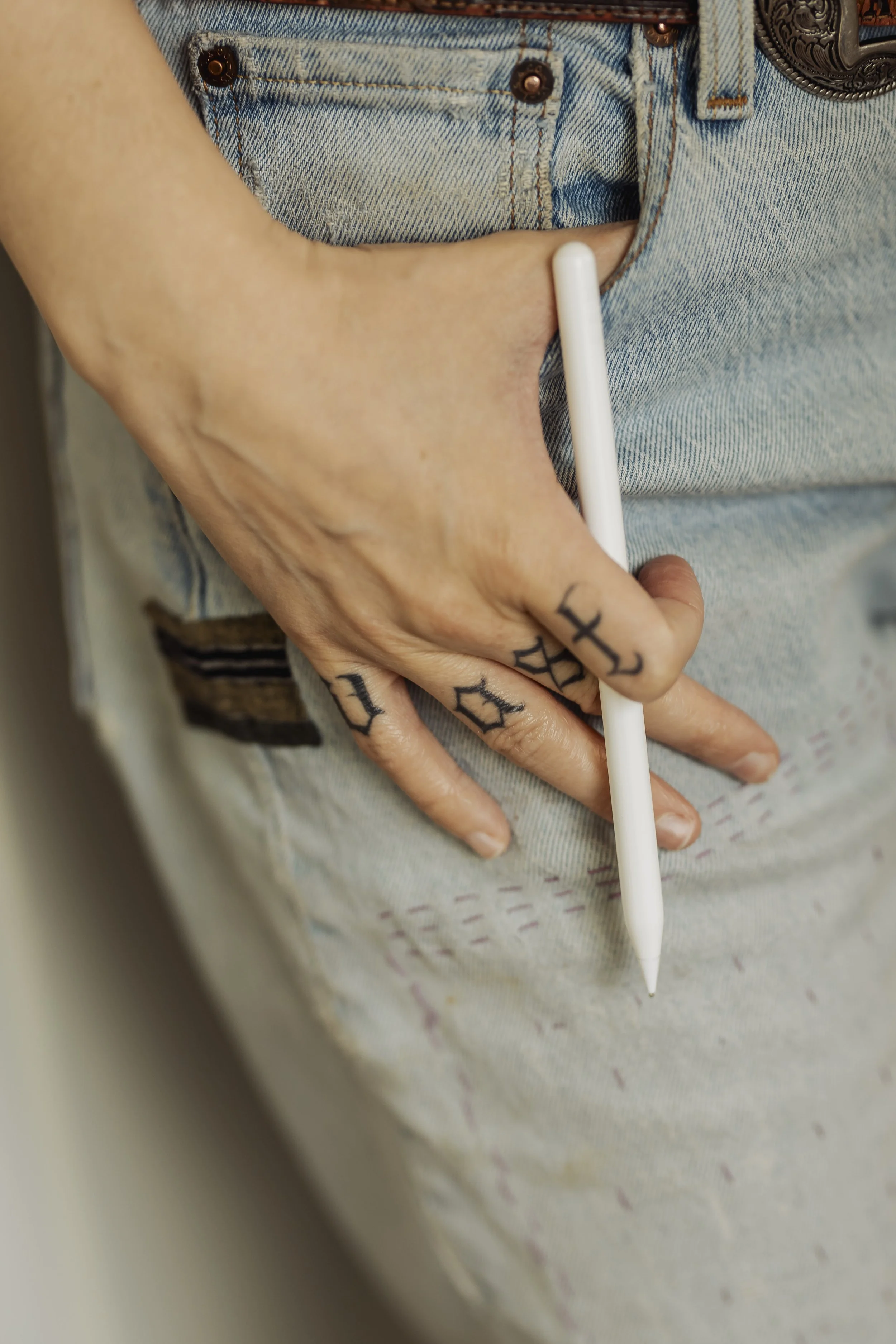 A person wearing light denim jeans has tattoos on their fingers and is holding a white stylus with their middle and ring fingers.