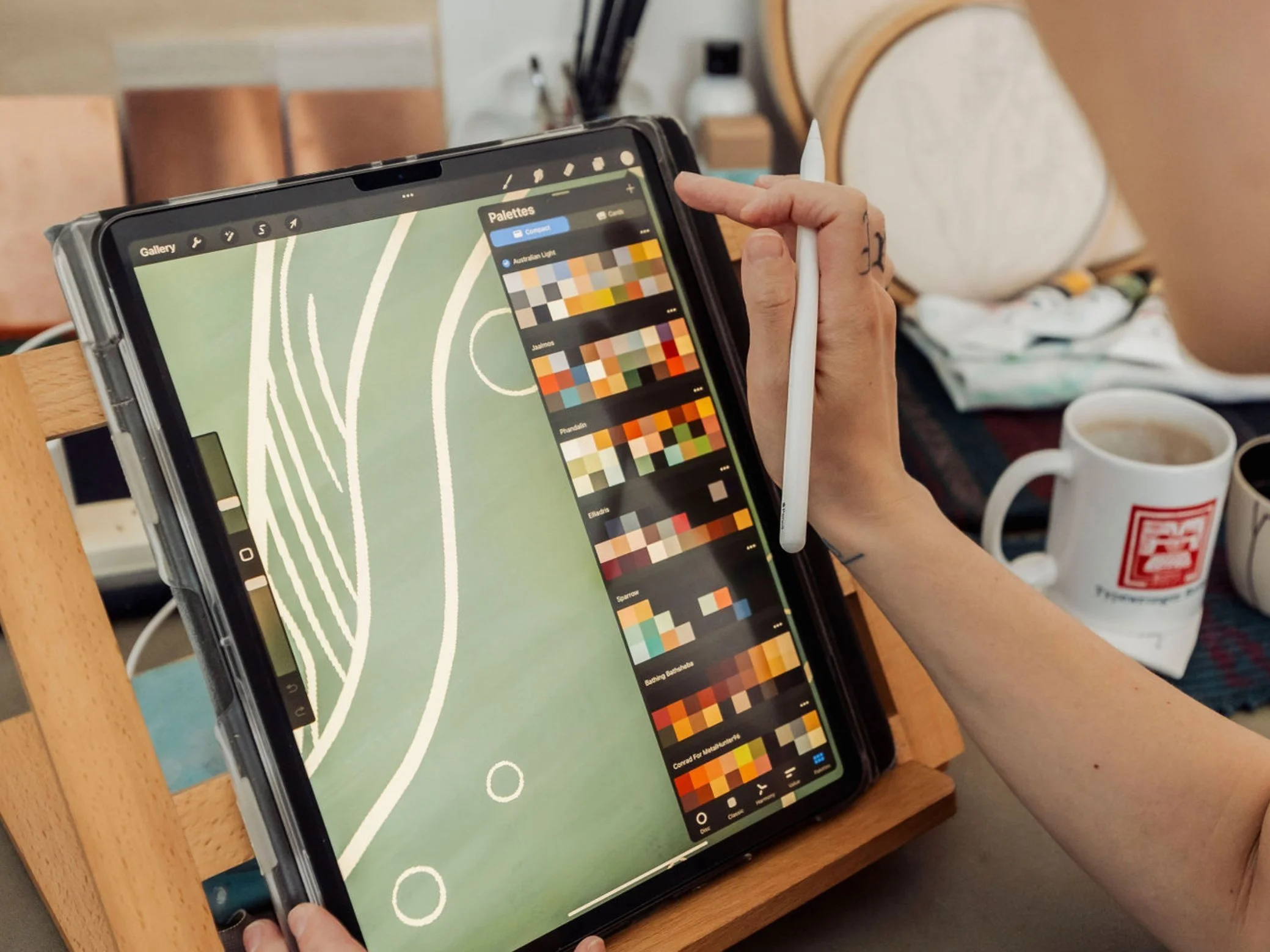 Person holding a tablet with a digital painting app open, displaying a design with white line art on a green background, sitting at a wooden table with coffee mugs and a plate in the background.