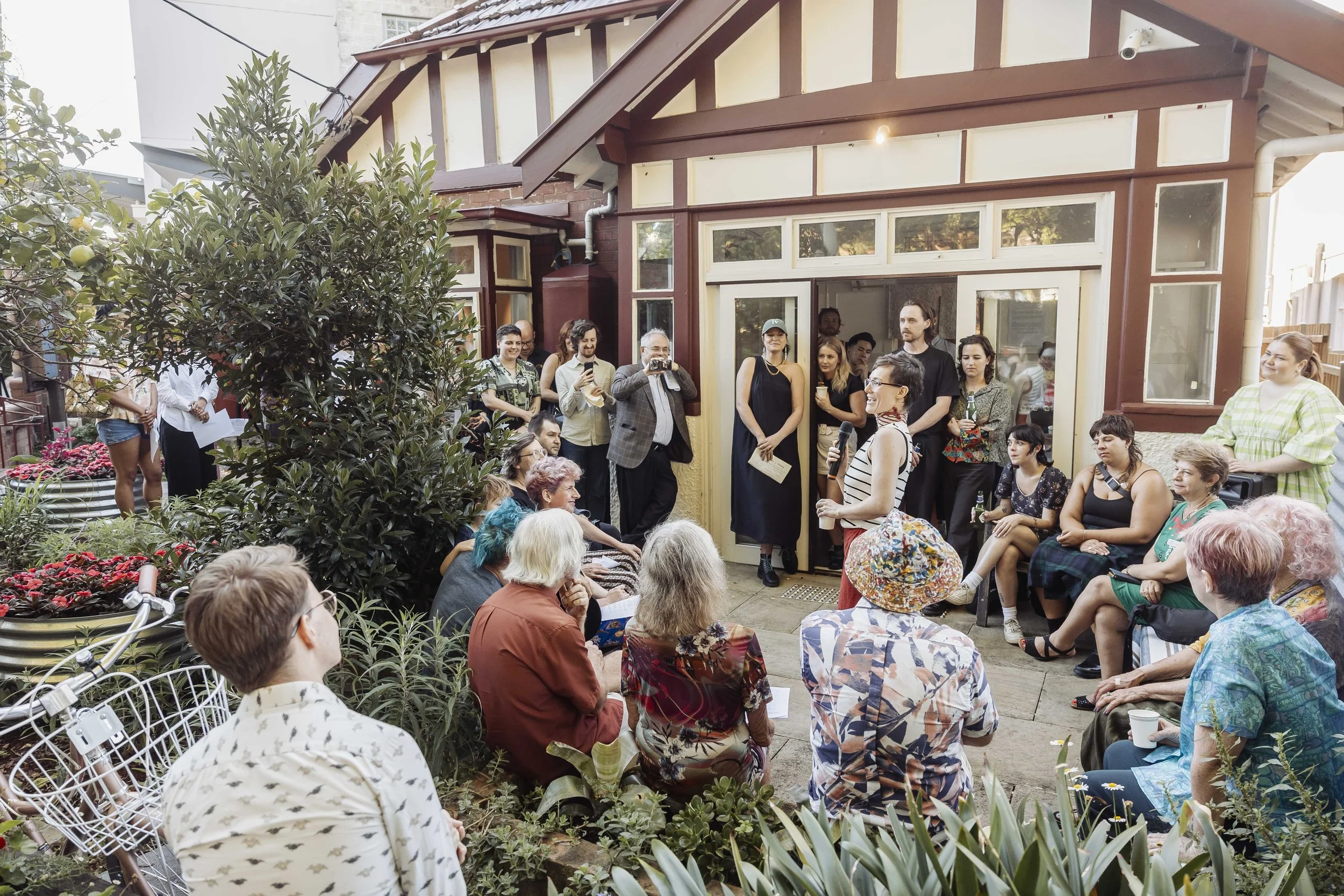 Holding a microphone in a crowded outdoor garden, a non-binary artist in a red wrap skirt addresses a crowd attending the opening night of an art exhibit at Barrett House Studios in Randwick, Sydney.