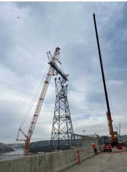 Construction workers using cranes to work on a tall metal power line tower along a waterfront.
