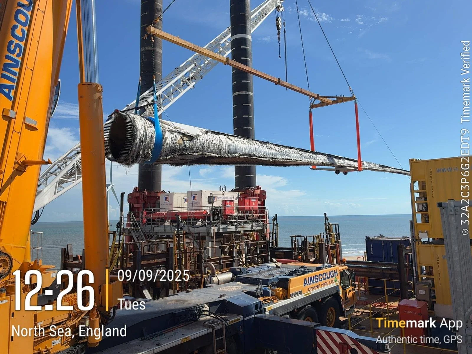 Offshore construction site on the North Sea, England, with a crane lowering a large, insulated pipeline over a platform near the ocean.